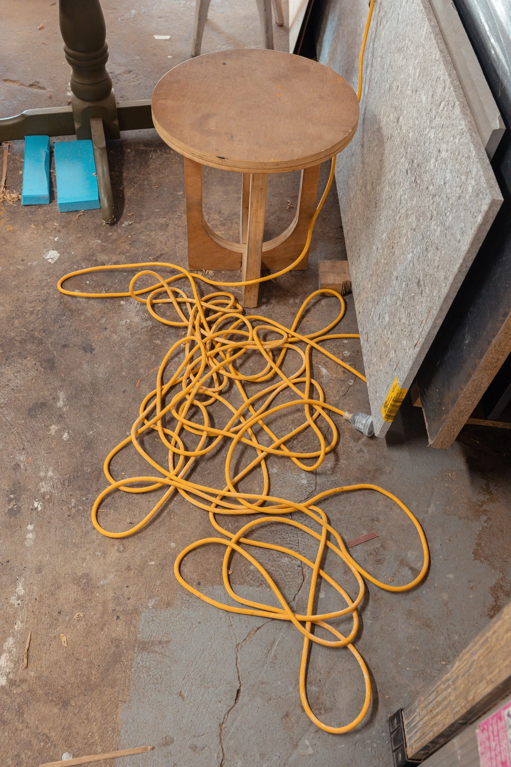 A wooden stool with a round seat and three legs on a concrete floor. An orange extension cord is tangled and spread on the floor in front of the stool, with a plug near the bottom right.