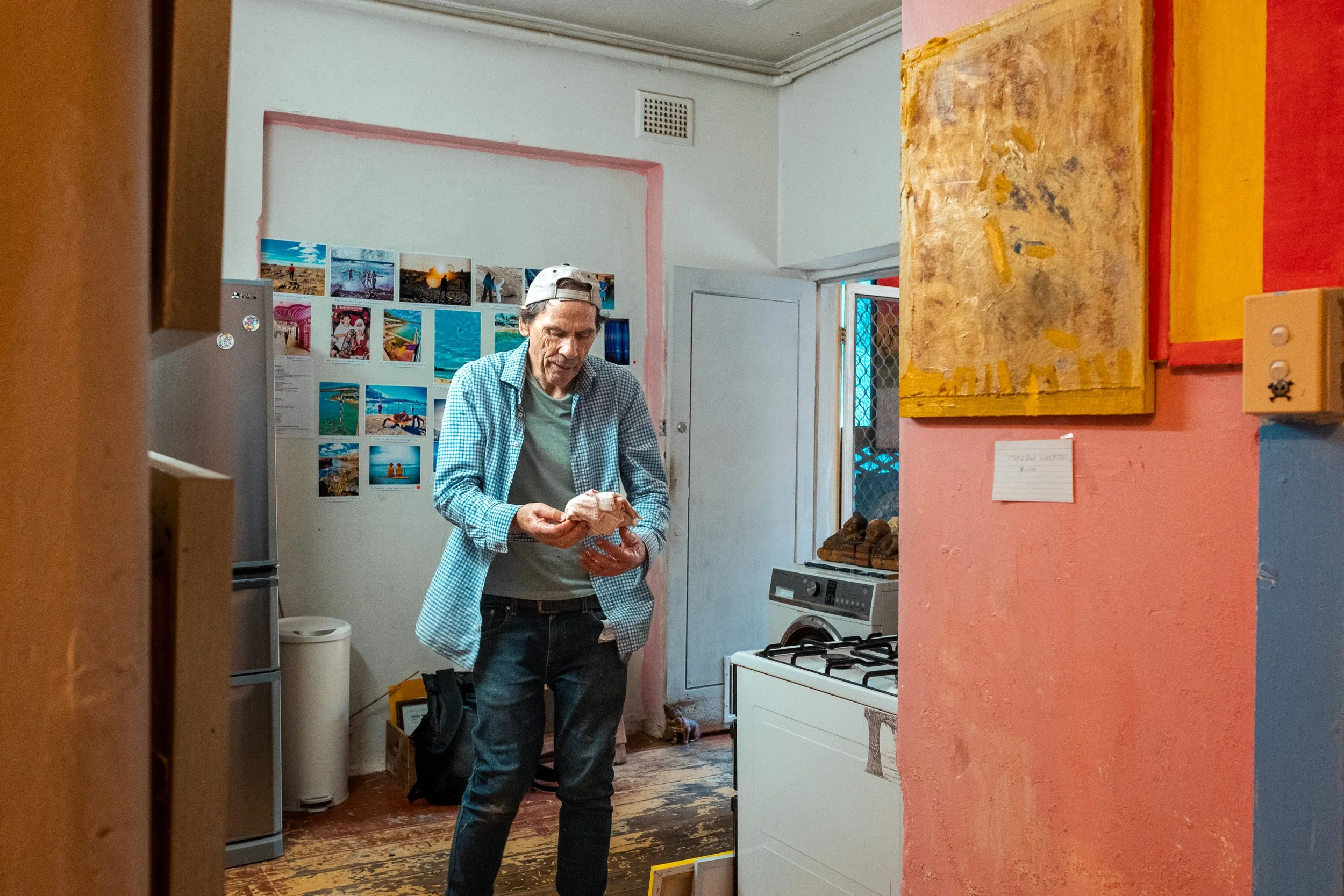 Man in casual clothing and baseball cap looking at a wrapped object inside a room with photos on the wall and colorful paintings on the walls.