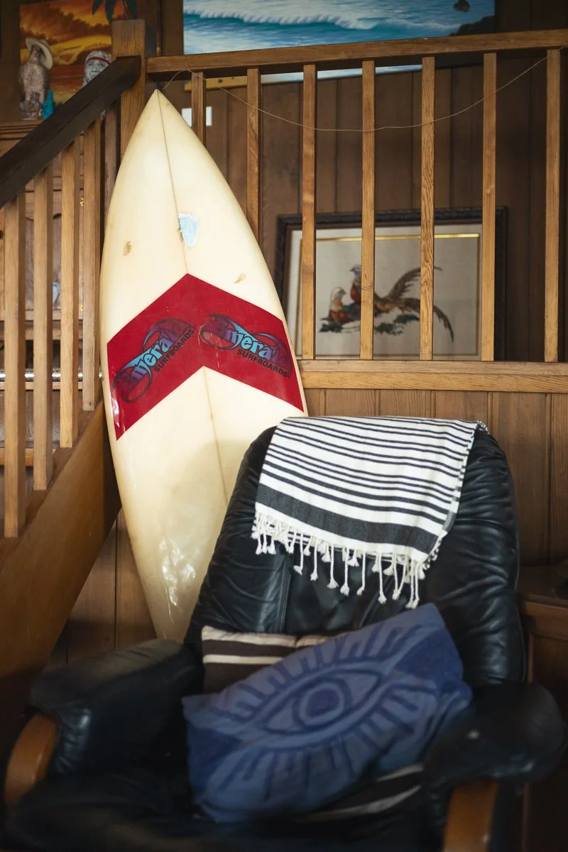 An indoor scene with a vintage surfboard leaning against a wooden railing and a black leather chair with a striped towel and a pillow on it, with wood-paneled walls and framed artwork in the background.