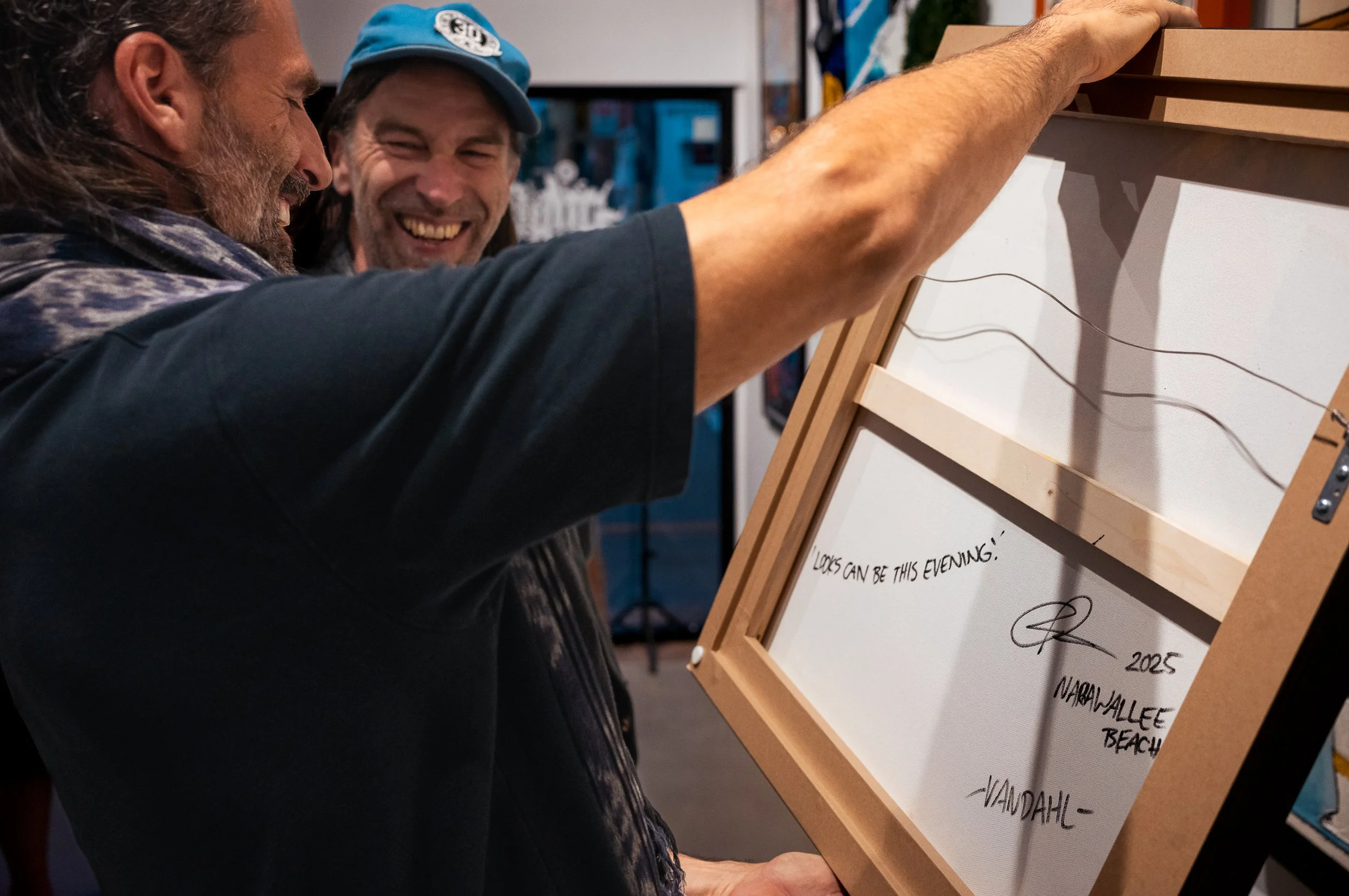 Two men smiling and looking at a framed artwork or signed print, which features handwritten text, a signature, and a date, in an indoor setting.