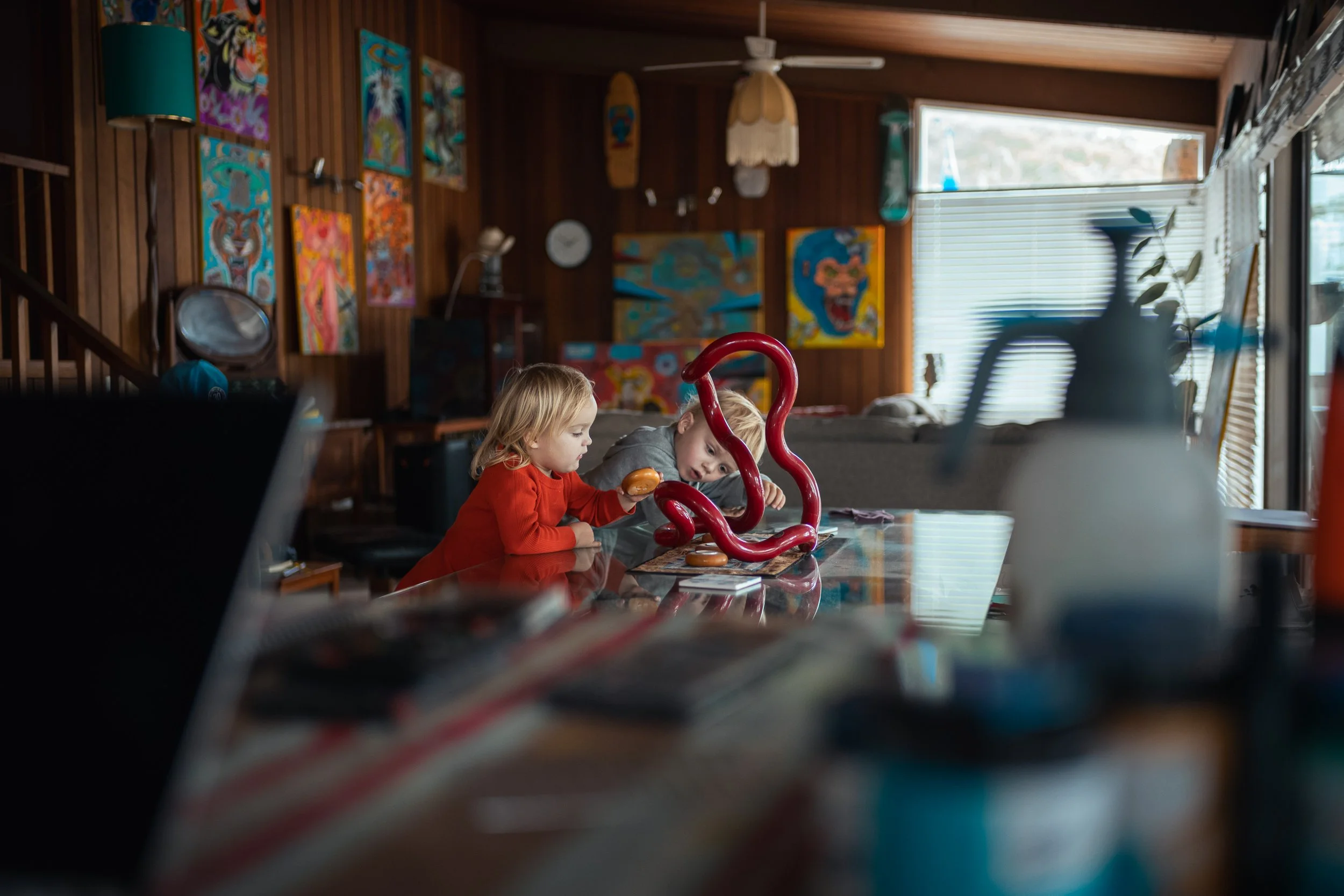 Two children, a girl with blonde hair in a red shirt and a boy with blonde hair in a gray shirt, sitting at a wooden table in a room with wooden walls and colorful artwork, including paintings and a skateboard, on the walls. The girl is holding a coo