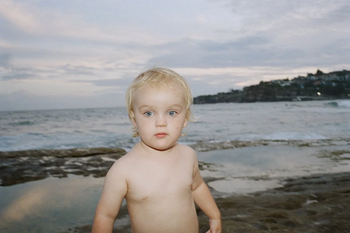 A young blonde-haired child with blue eyes standing shirtless on a beach, with ocean waves and a cloudy sky in the background.