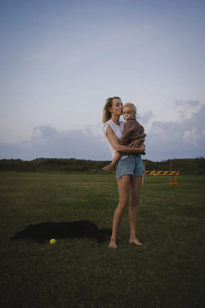 A woman holding a child in an outdoor grassy area with cloudy sky, a tennis ball and a black dog laying on the ground nearby, and a distant orange safety barrier.