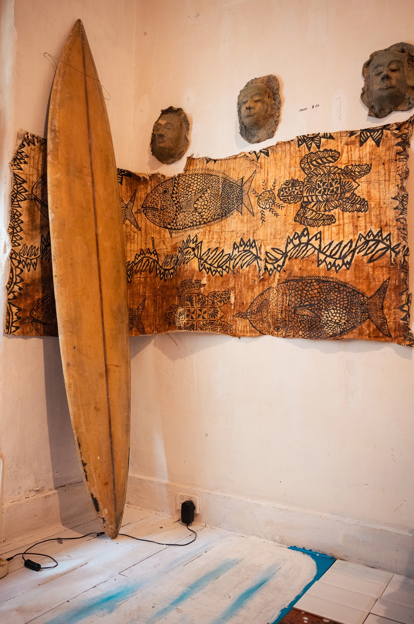 A wooden surfboard leaning against a wall, a decorative tapestry with fish and leaf patterns, and three tribal-style mask sculptures mounted on the wall above.