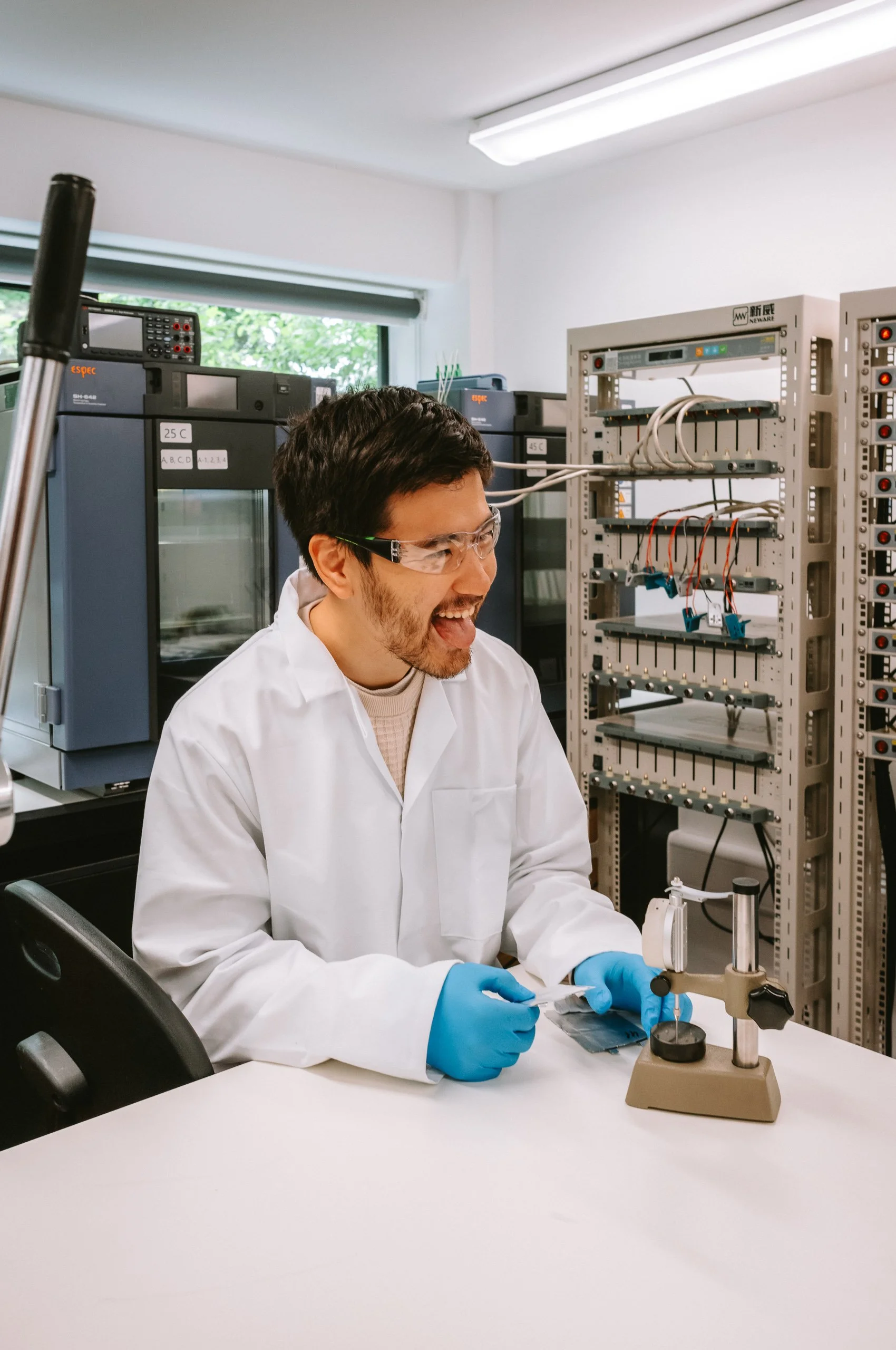 A scientist in safety glasses, blue gloves, and a lab coat working with a microscope in a laboratory with scientific equipment and electrical equipment in the background.