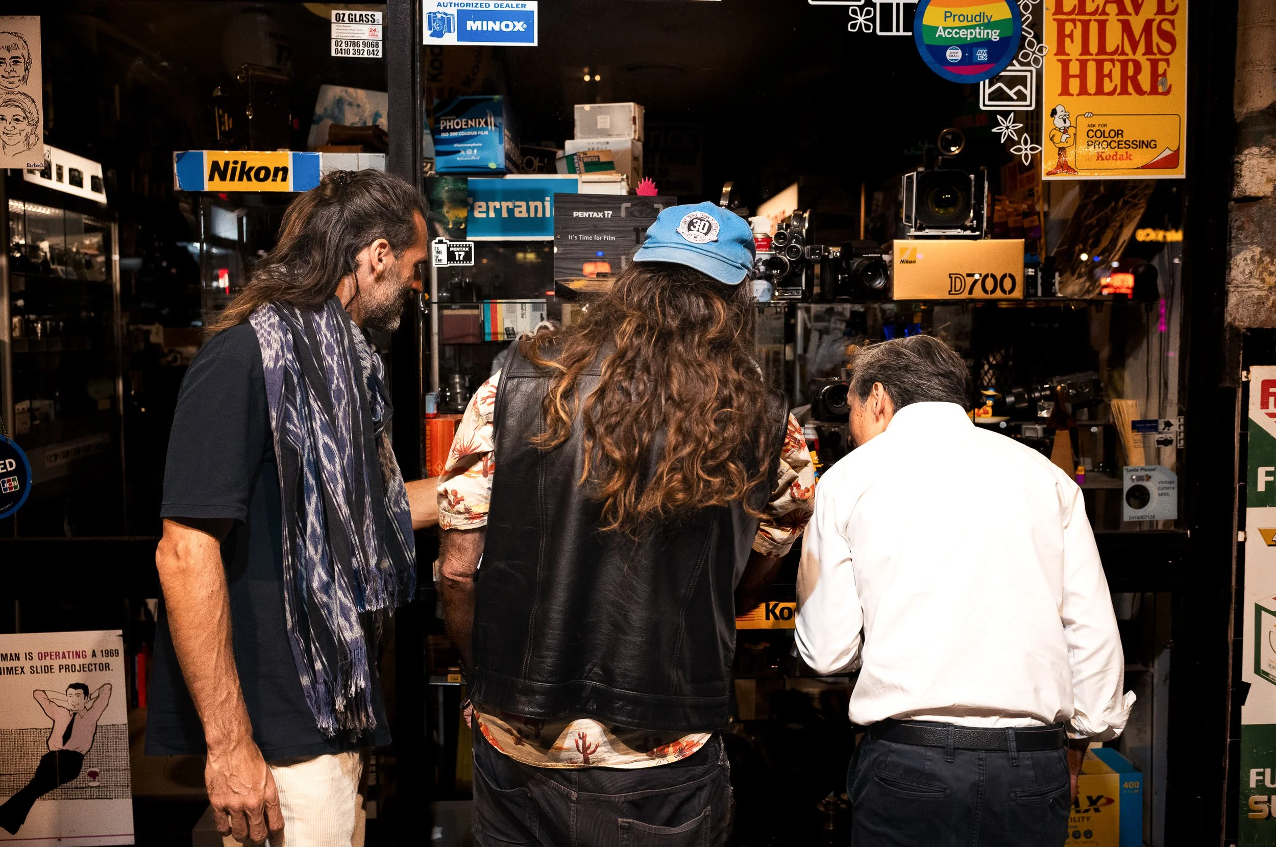 Three men with long hair are looking at camera equipment inside a store that sells photo and video gear. One man is wearing a black shirt with a blue and white scarf, another is in a black leather vest with a light blue cap, and the third is dressed 