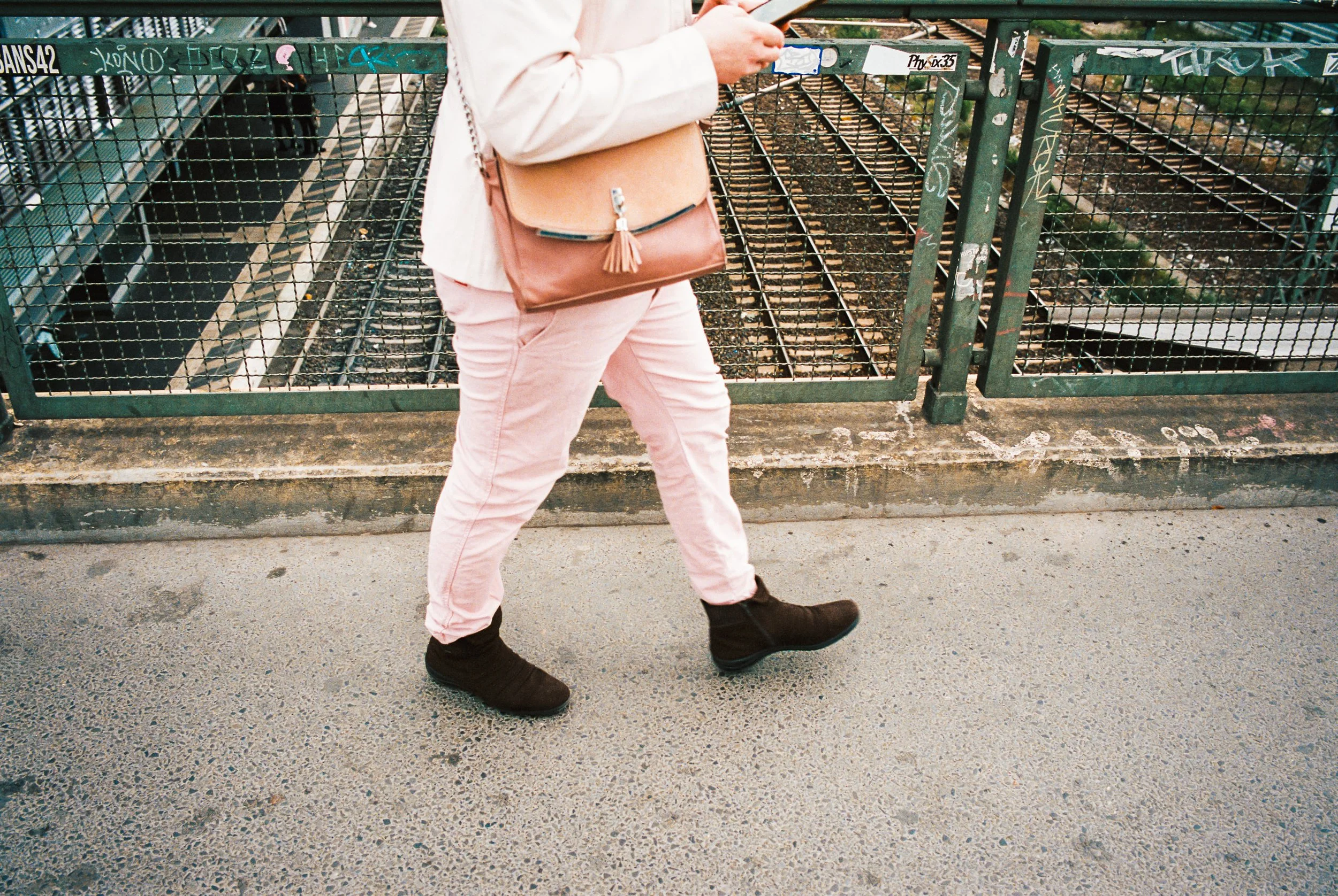 Person walking on sidewalk next to a metal fence with graffiti, wearing pink pants, a beige jacket, black shoes, and carrying a tan purse.