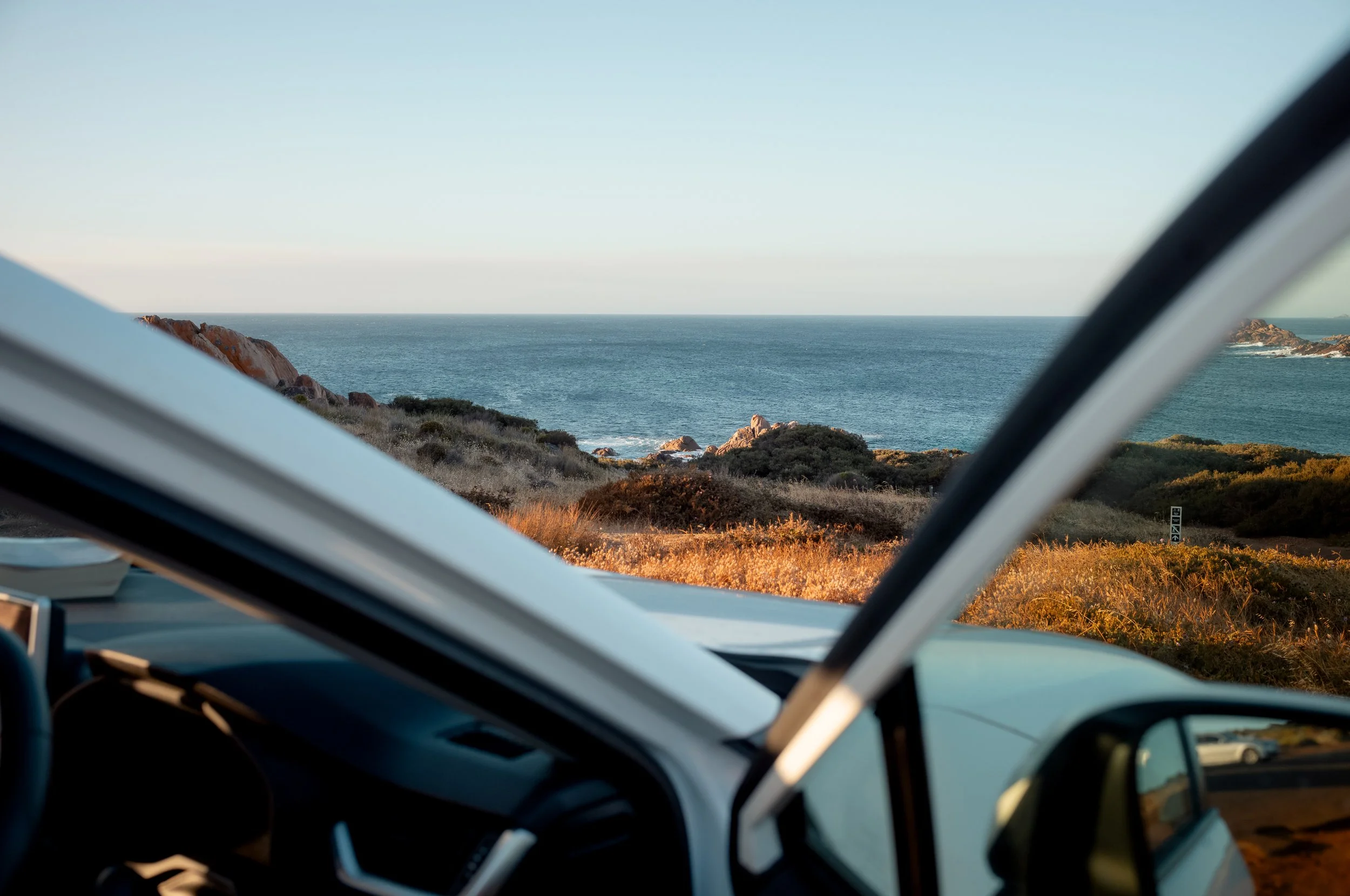 View of the ocean with rocky cliffs and sandy beach, seen through the windshield of a vehicle parked on a coastal road during sunset.