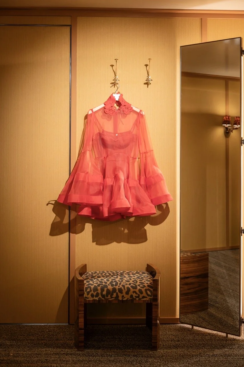 A red dress hanging on a wall hook, with a leopard print stool below it, in a room with wood panel walls and a full-length mirror on the right.