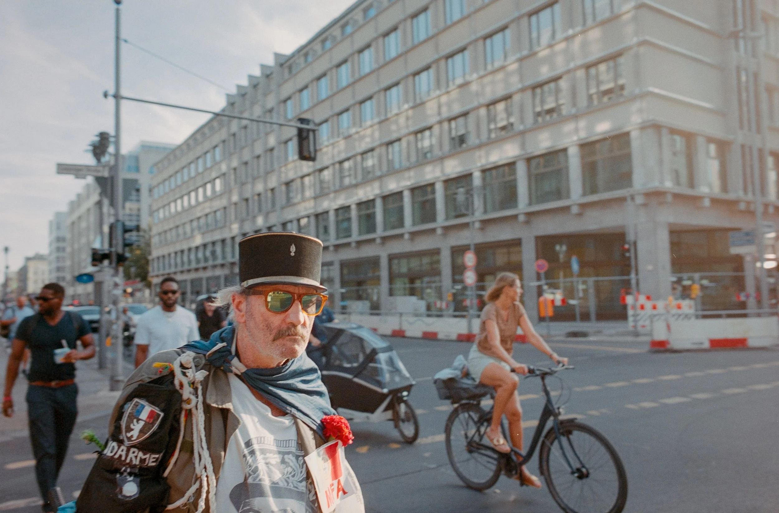 A man wearing a black top hat, sunglasses, and a patterned scarf walks on a city street with other pedestrians and a cyclist in the background during daytime.