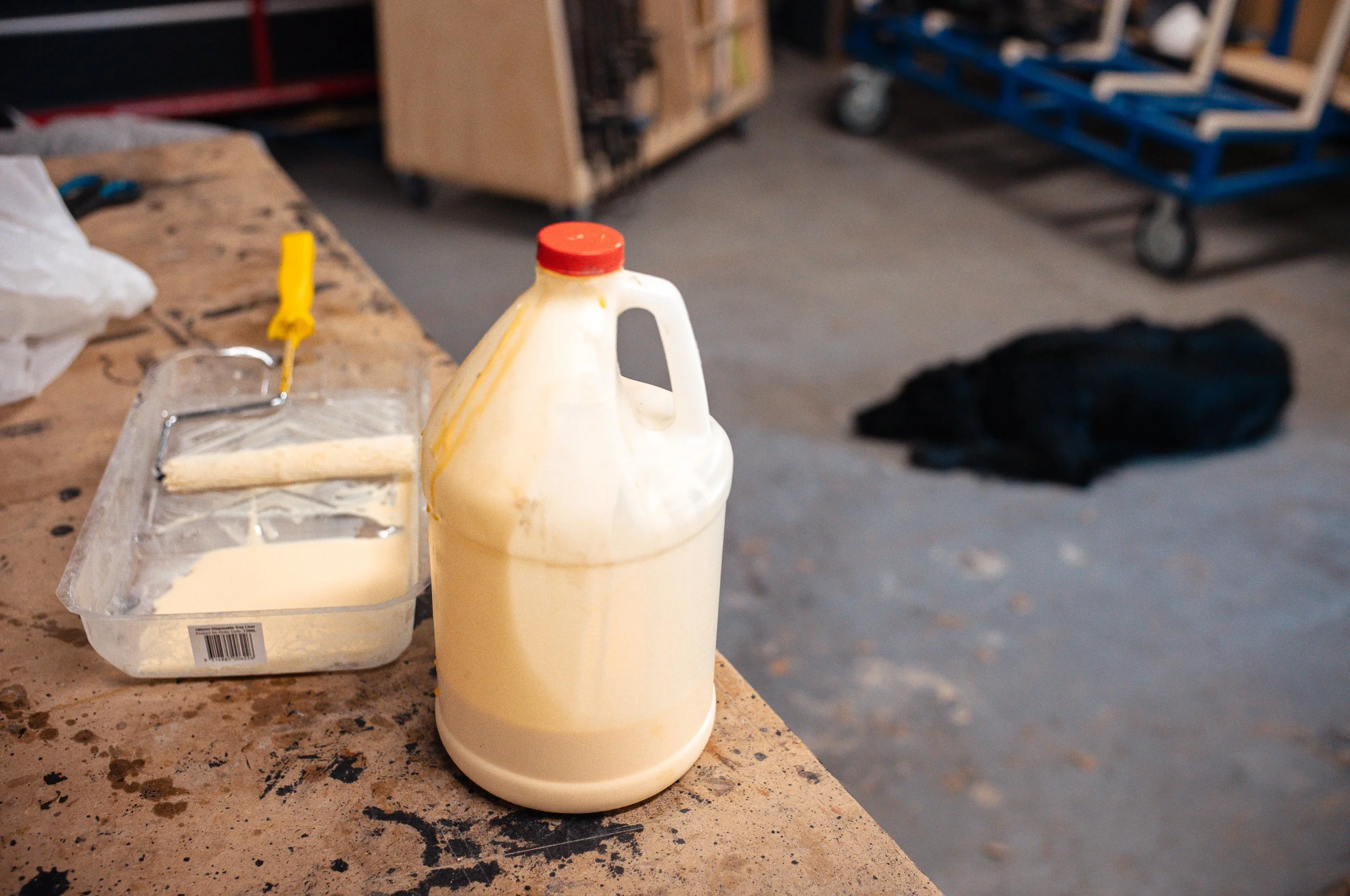 A cardboard workbench with a large plastic jug of yellow paint, a paint roller, and a paint tray with some leftover paint. In the background, a blue cart with wheels and a black backpack on the floor.