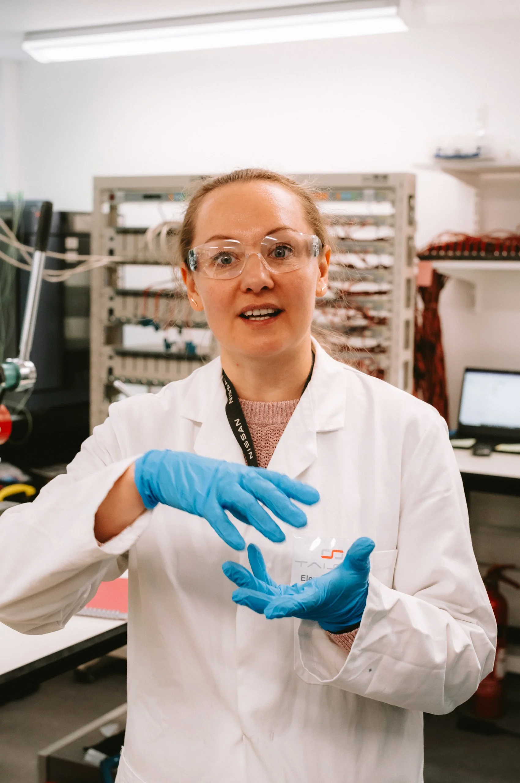 A woman in a lab coat and safety goggles wearing blue gloves, working in a laboratory.