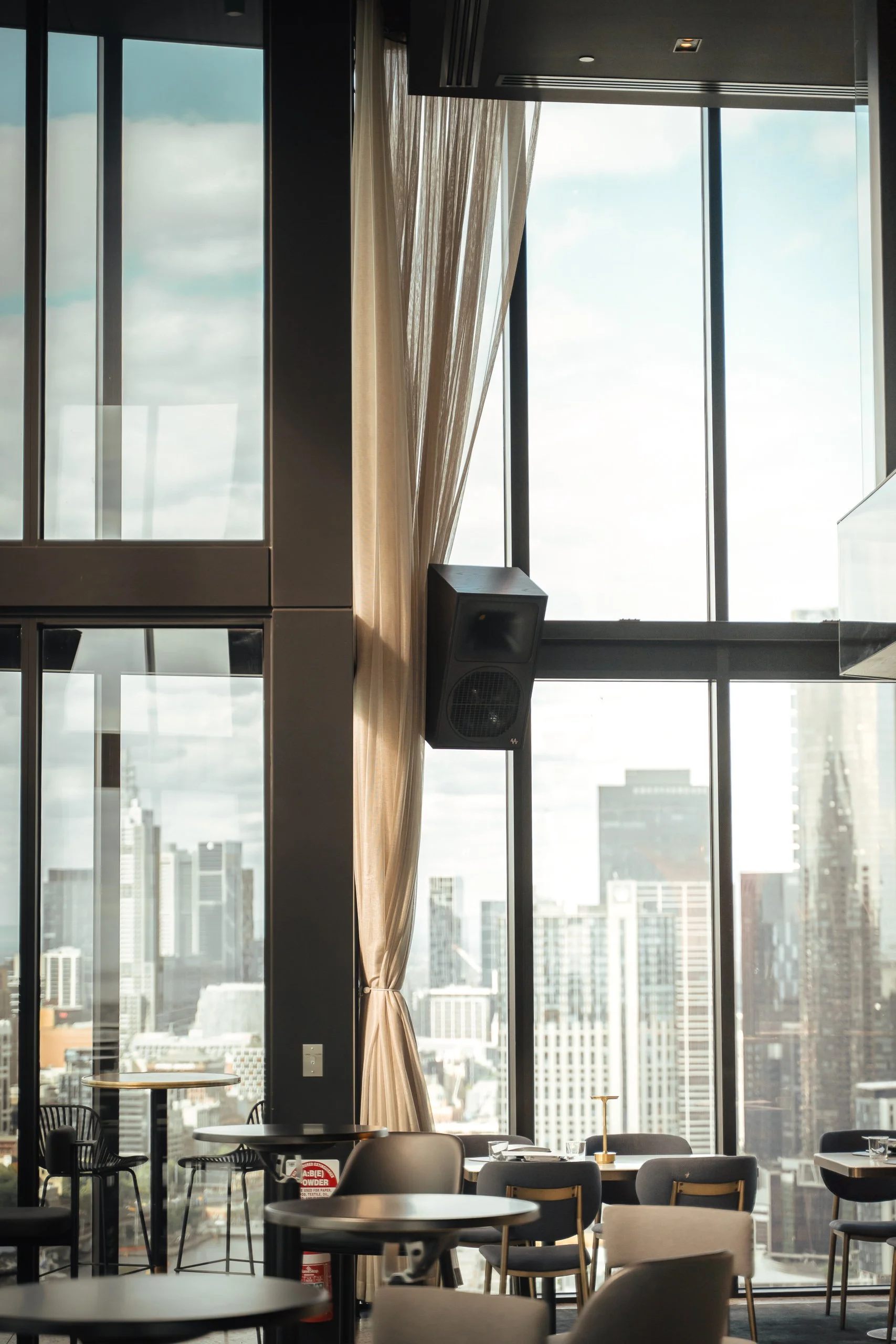 Interior of a modern high-rise restaurant or lounge with large windows showcasing a city skyline, featuring round tables, modern chairs, and a speaker mounted near the window.