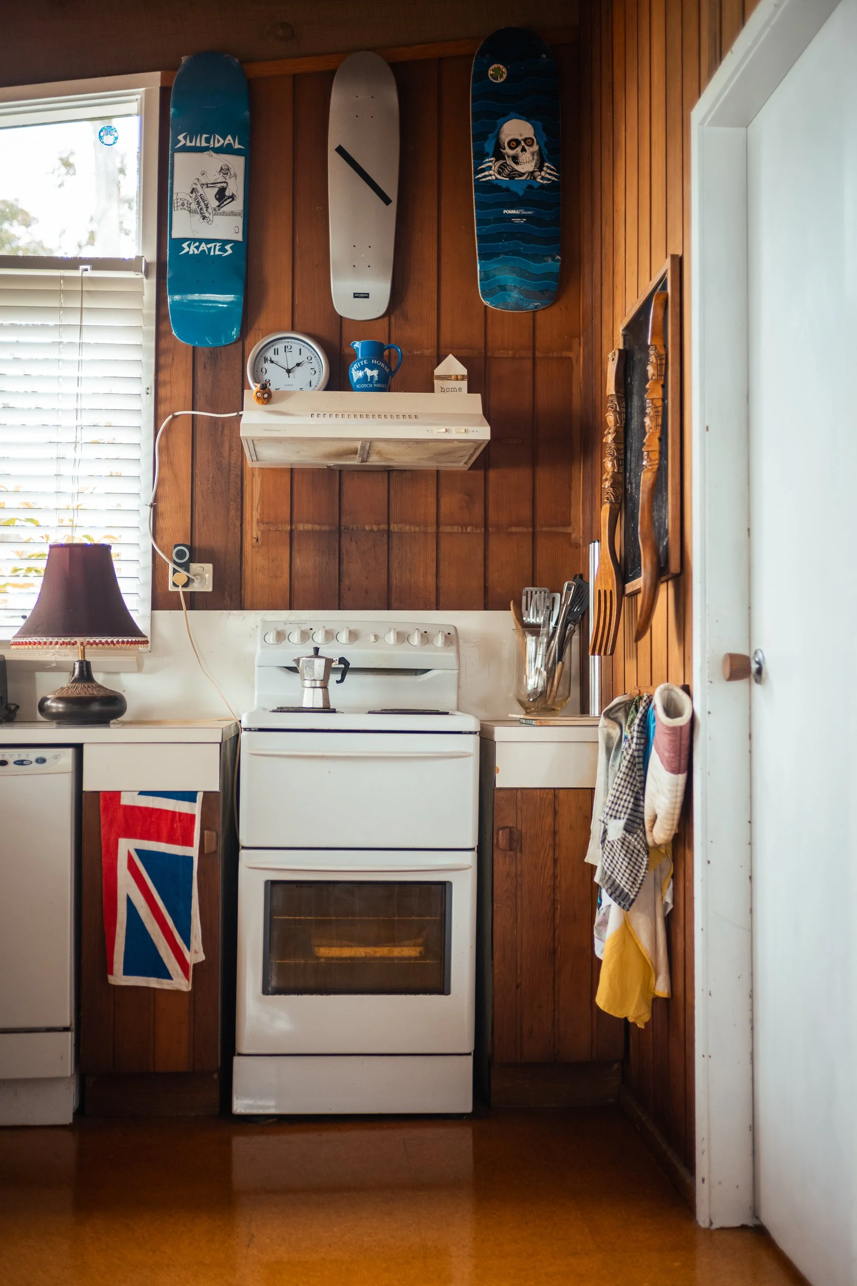 View of a vintage kitchen with wood paneling, white stove, and kitchen utensils. Decor includes skateboard decks on the wall, a clock, and a blue mug, with a small window and a lamp on the left.
