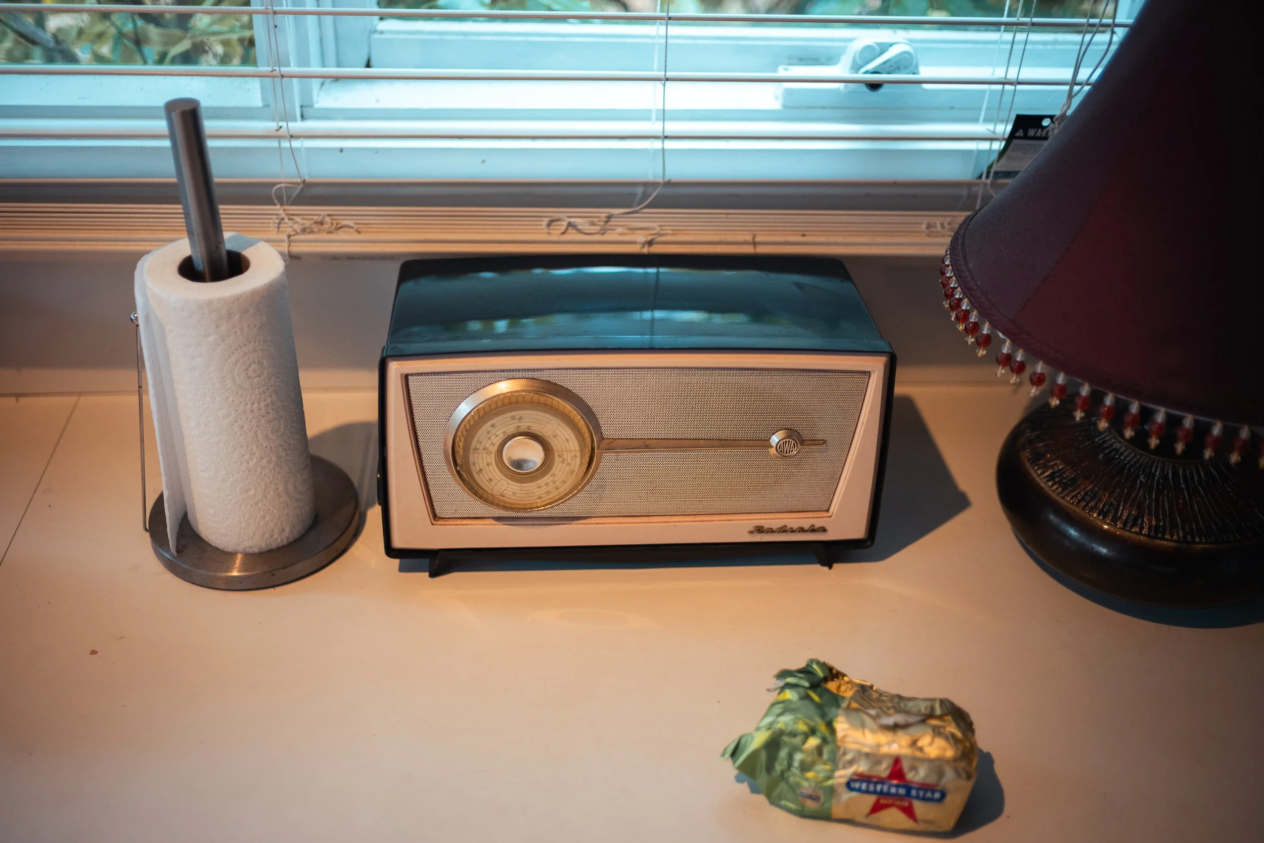 A vintage radio, a paper towel roll on a metal stand, and a bag of shredded cheese on a white countertop next to a window with blinds.
