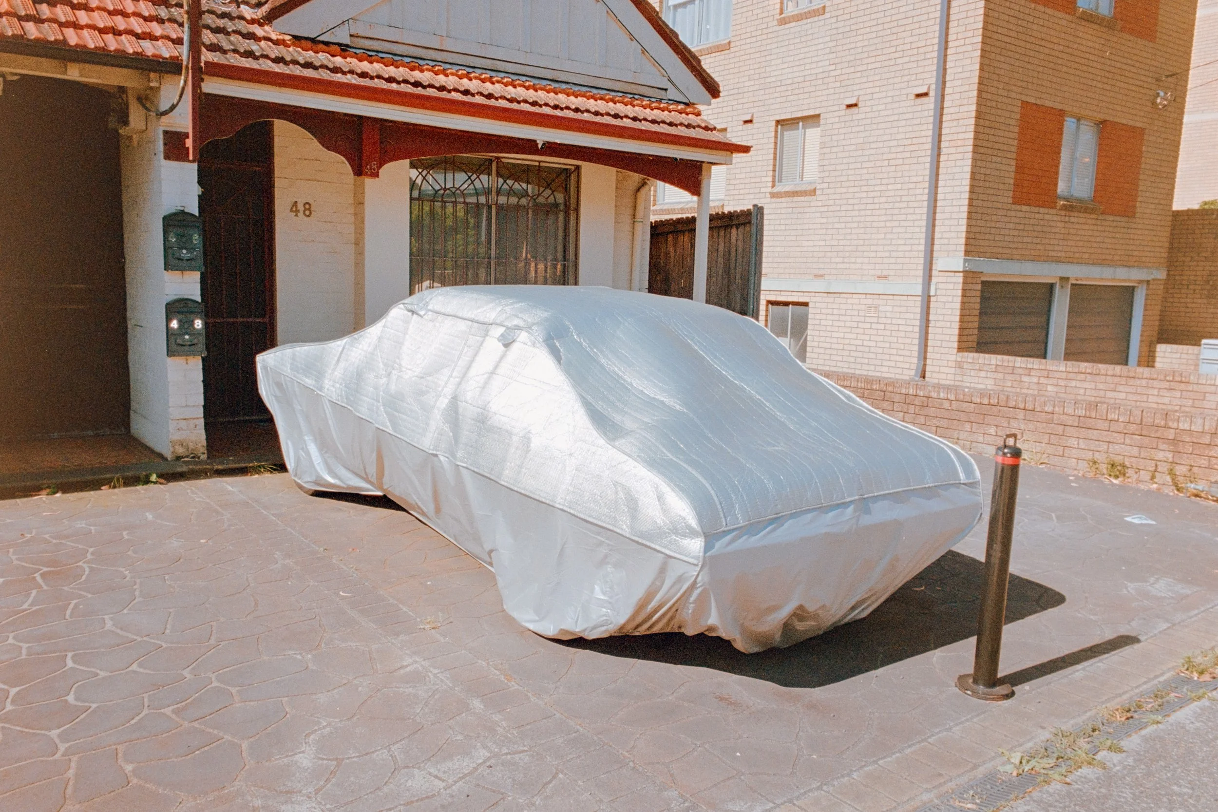 A car covered with a silver protective cover parked on a driveway in front of a house with a red tiled roof and brick walls.