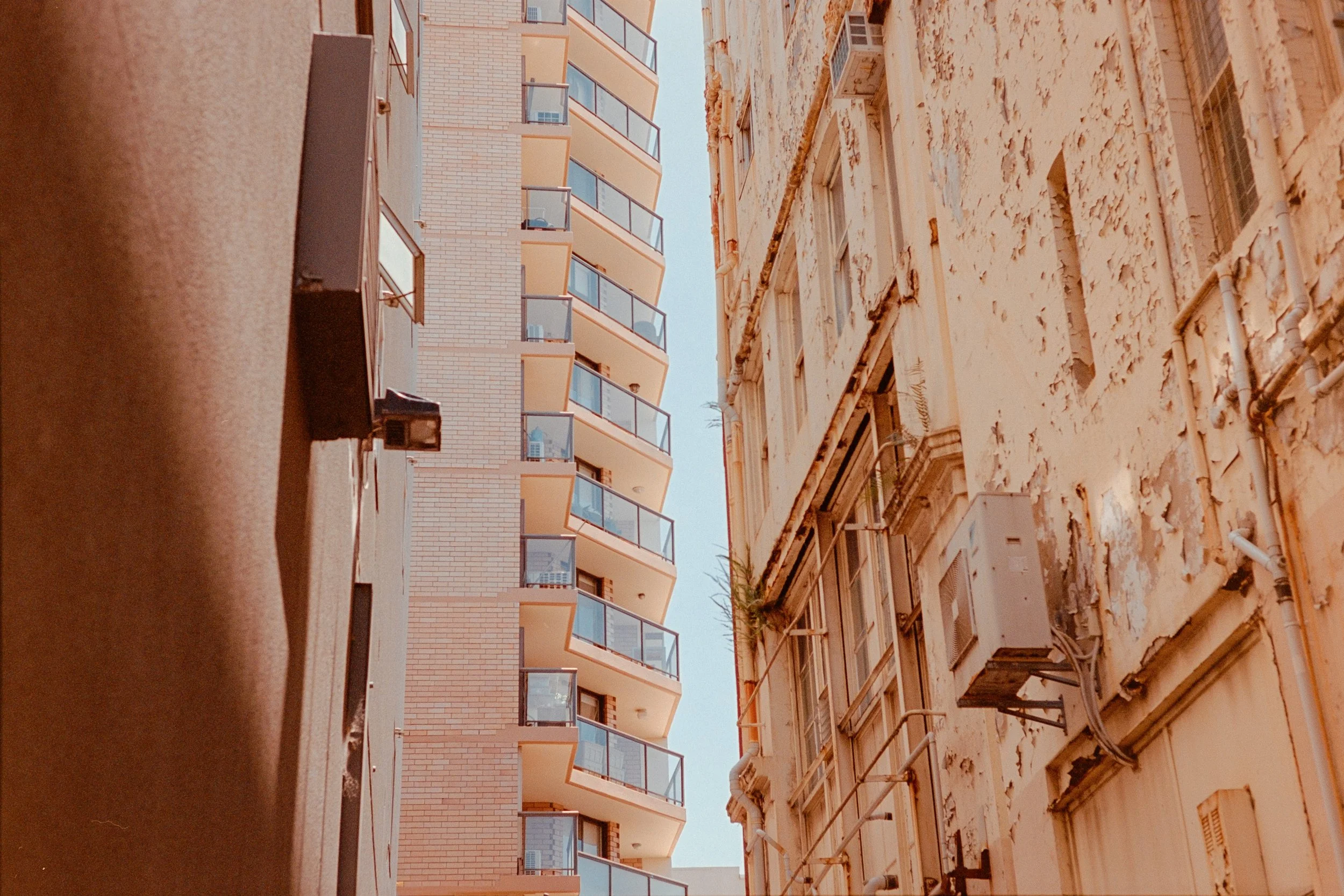 Looking up between two buildings, one modern with balconies and one old with peeling paint, against a clear blue sky.