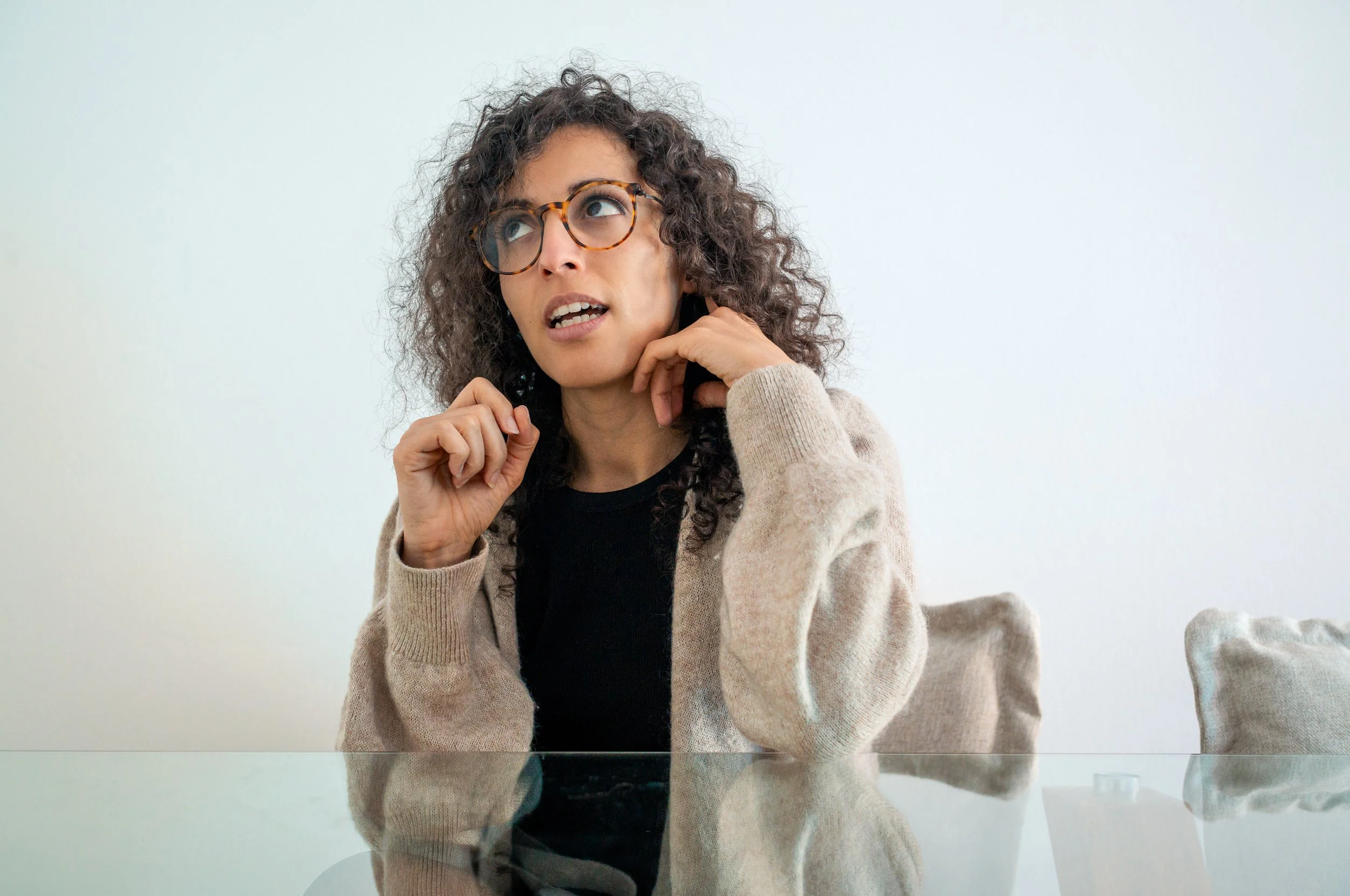 A woman with curly hair and glasses sitting at a glass table, talking on a cellphone.