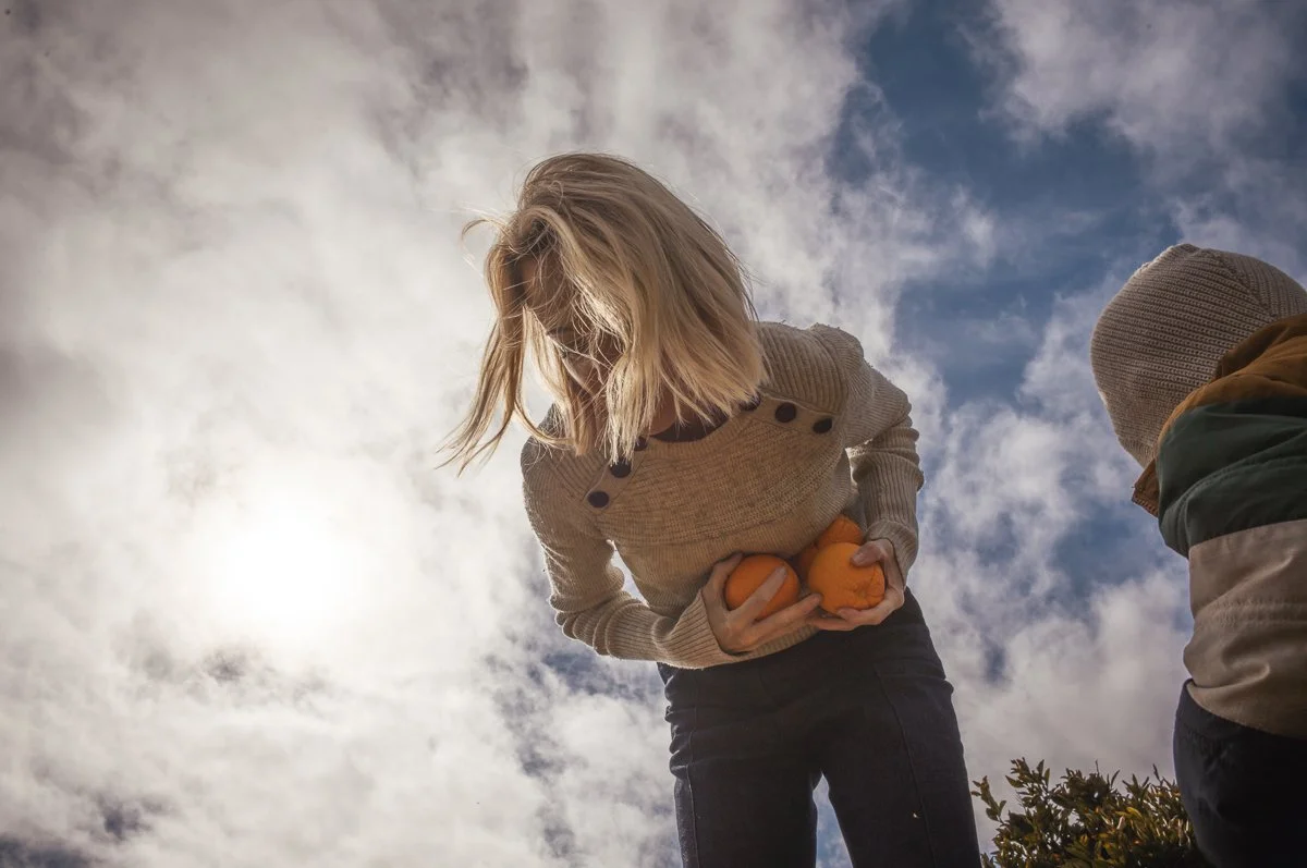A woman holds three small pumpkins outside on a cloudy day, with a young boy nearby looking at the pumpkins.