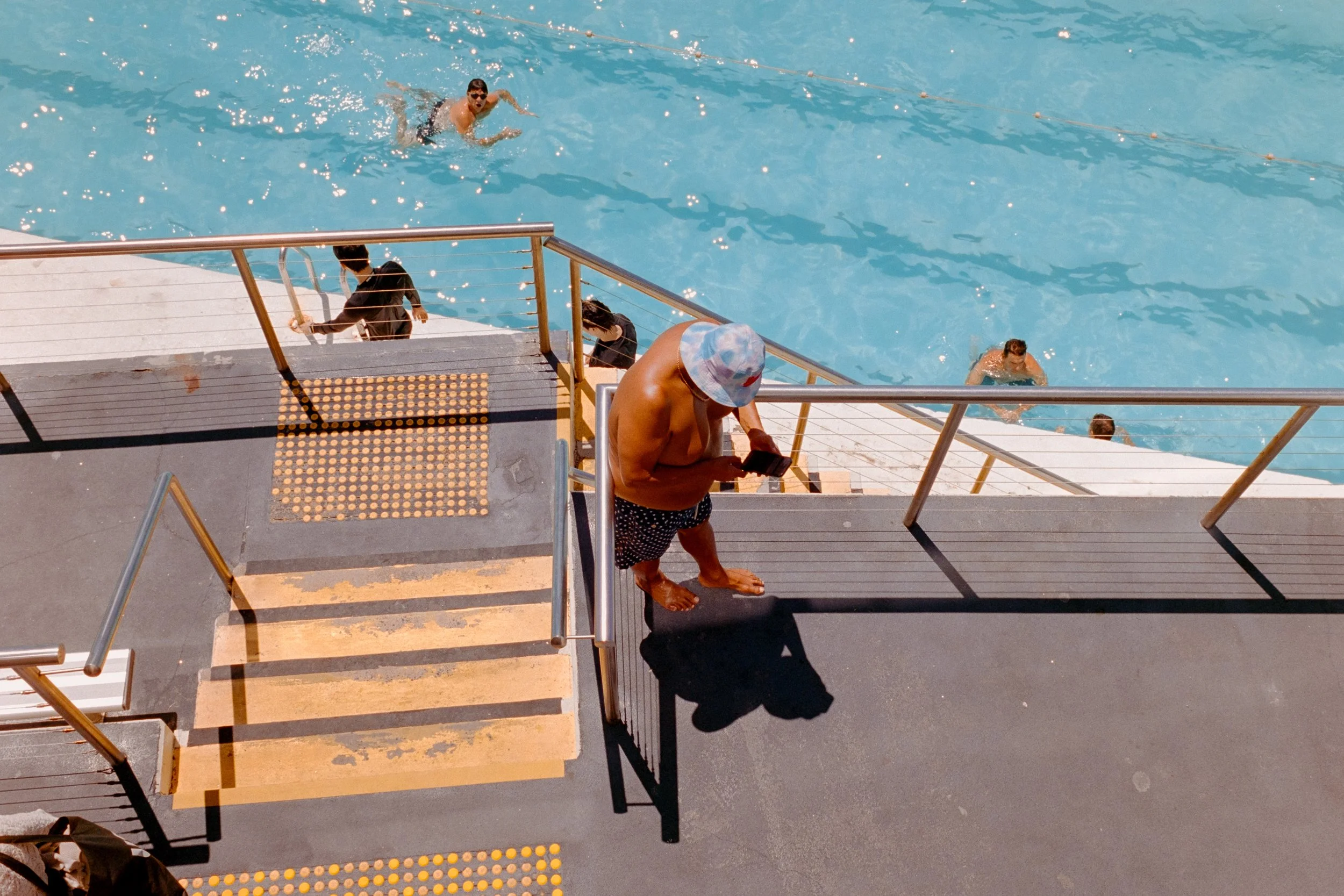 A person in swim trunks and a bucket hat standing on a pool deck, looking at their phone, while other people swim and rest in the pool.