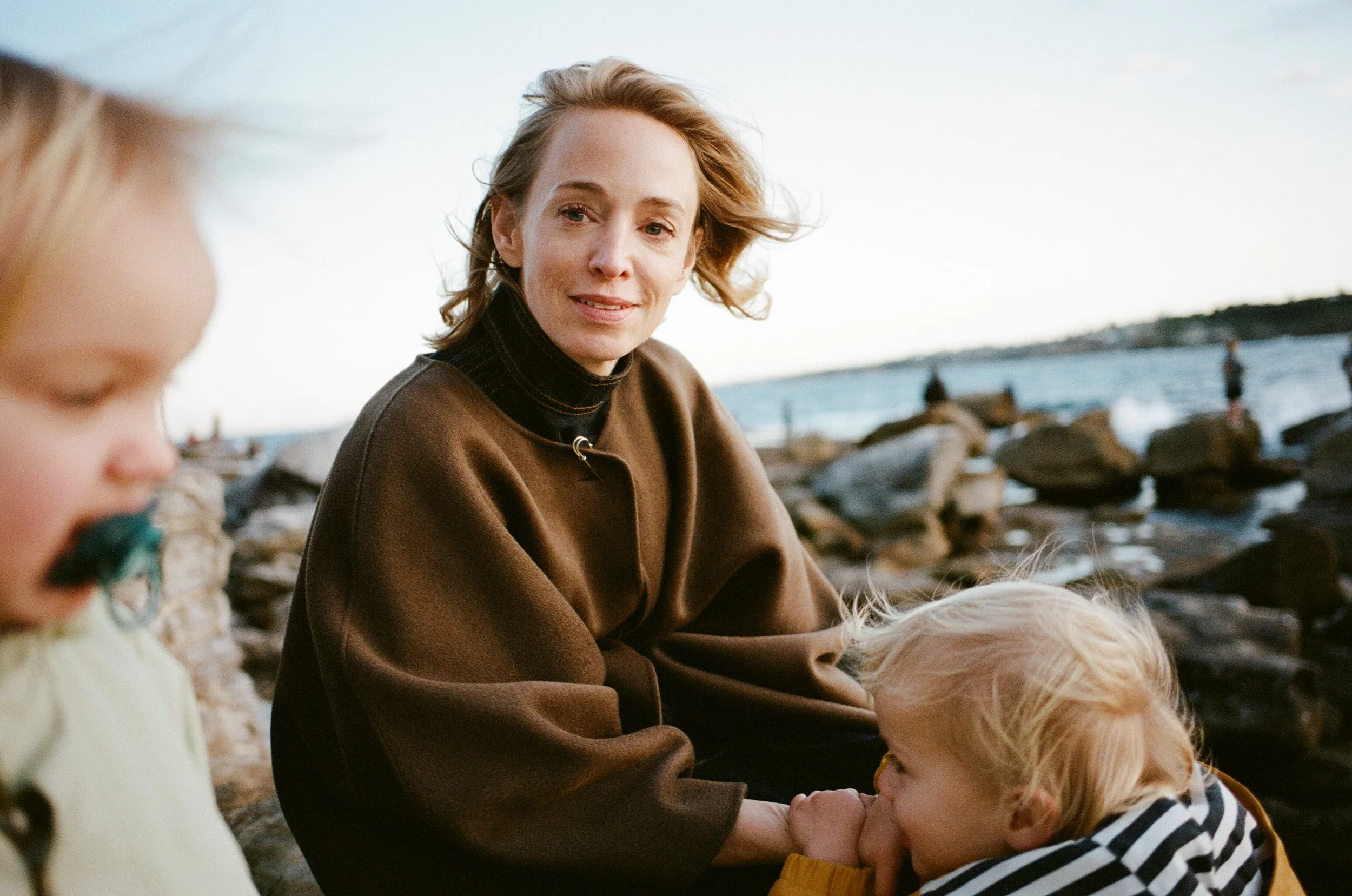A woman sitting on rocks at the beach, with two children, one with blond hair and a striped shirt, and the other with dark hair and pacifier, during sunset or late afternoon.