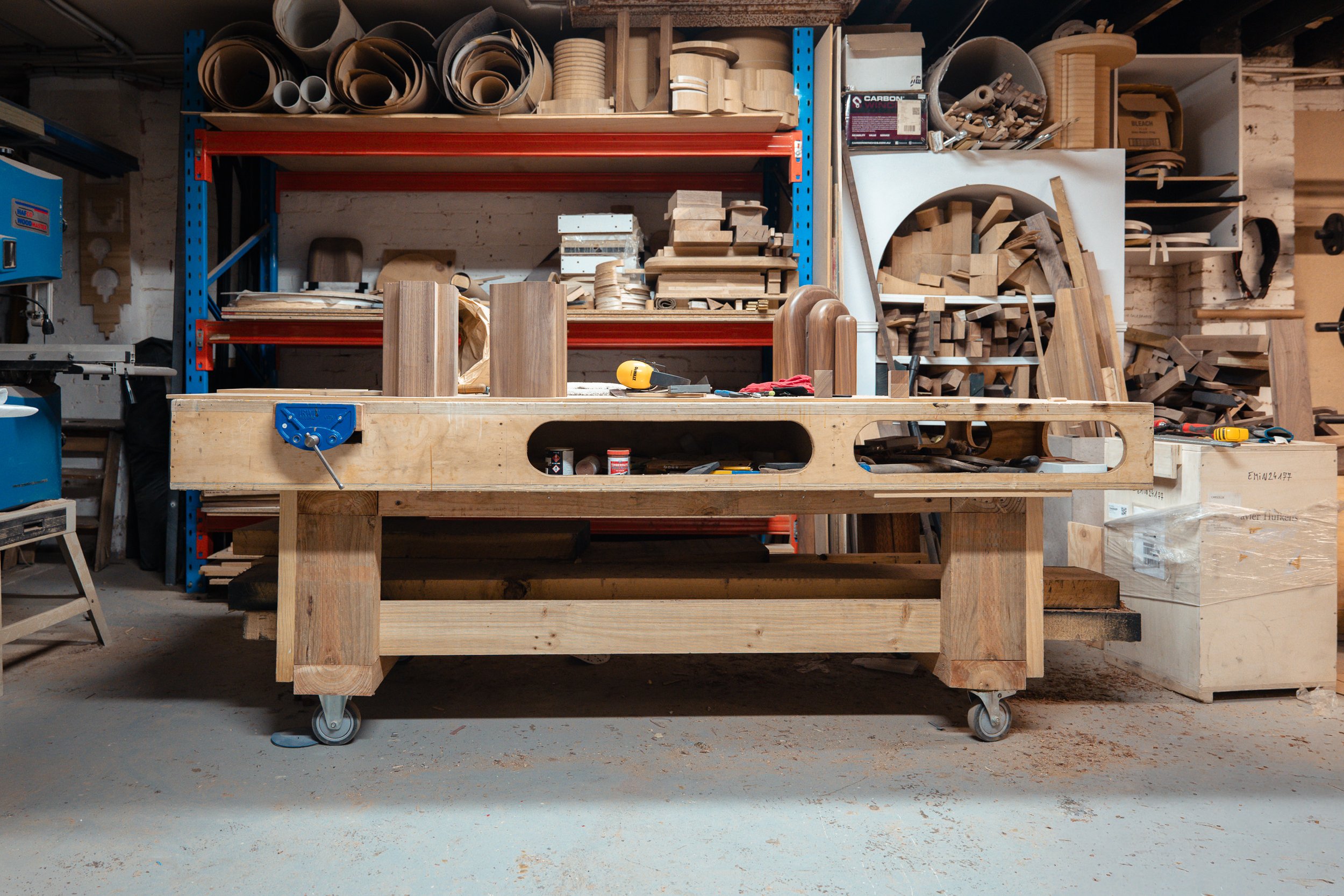 Woodworking workshop with a workbench, various wooden pieces, tools, and shelves filled with supplies.
