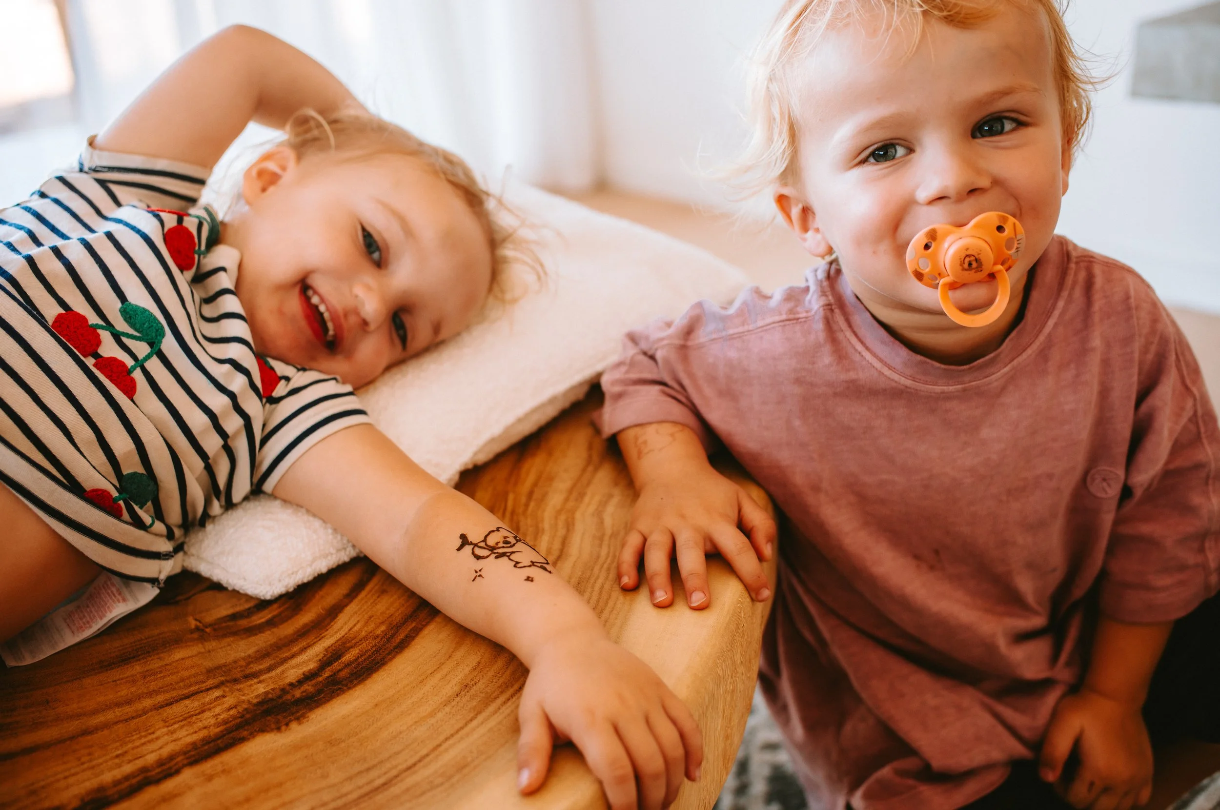 Two young children, a girl lying on a bed with a tattoo on her arm and a boy sitting beside her with a pacifier, smiling at the camera.