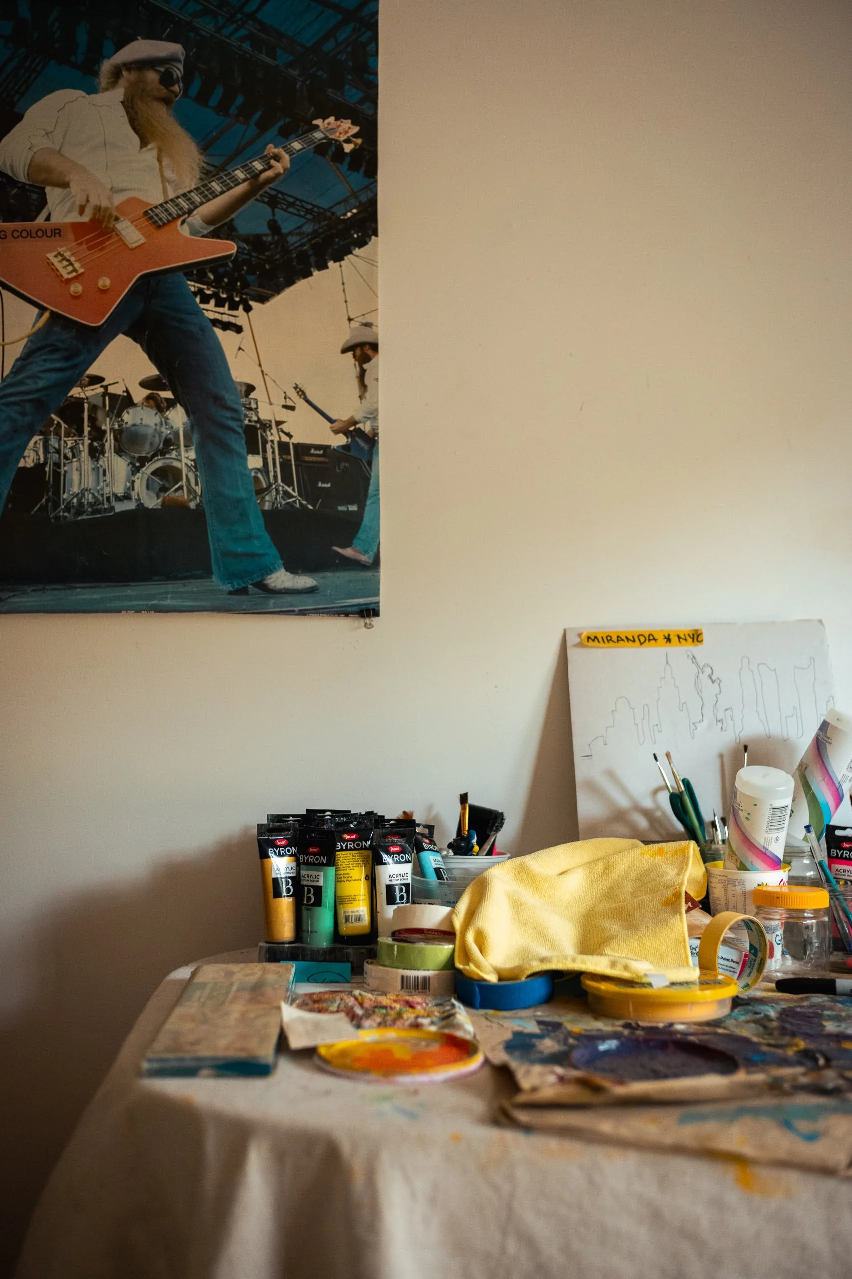 Artist's workspace with paints, brushes, and supplies on a table, and a white canvas with an outline of a city skyline and the Statue of Liberty, labeled 'MIRANDA # NYC'. In the background, a poster of a band performing on stage.