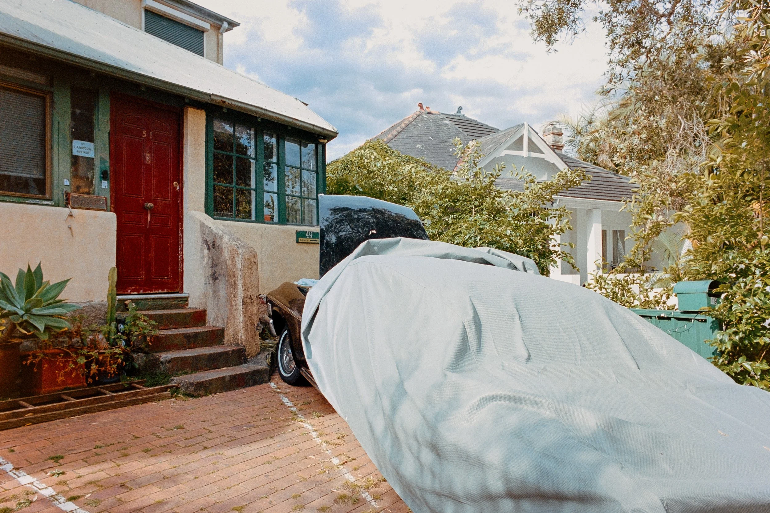 A house with a red front door, set on a brick sidewalk with steps leading up. A car parked in front, partially covered with a white tarp, and lush green trees surround the property.