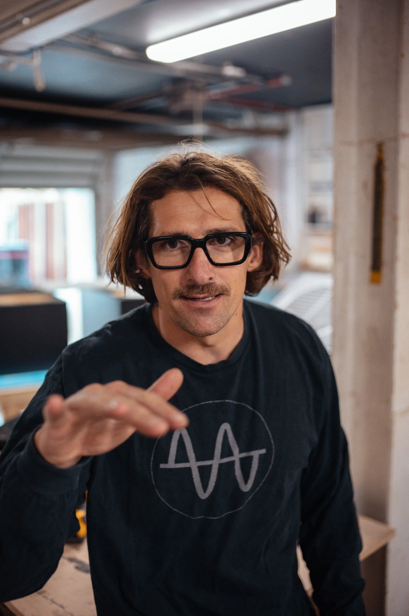 Man with brown hair, glasses, and mustache gesturing towards the camera, wearing a black shirt with a white wave-like symbol.