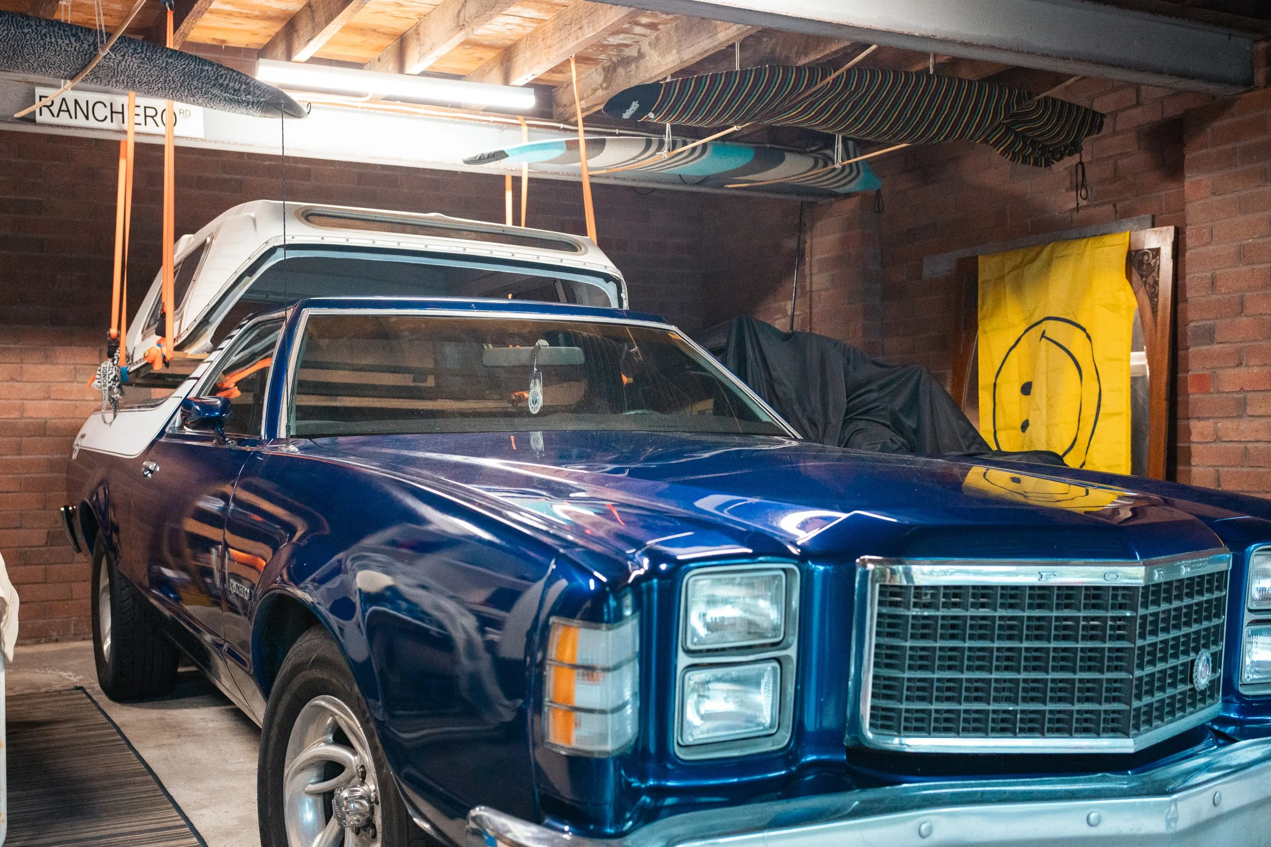 A blue vintage car parked inside a garage, with a white roof rack on top, and surfboards stored on the ceiling. There's a yellow flag with a smiley face on the wall.