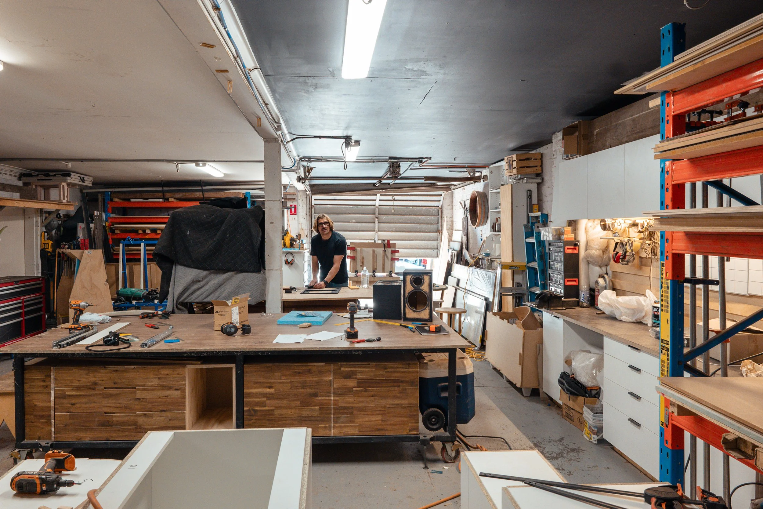 A man working in a woodworking workshop filled with tools, materials, and storage shelves.