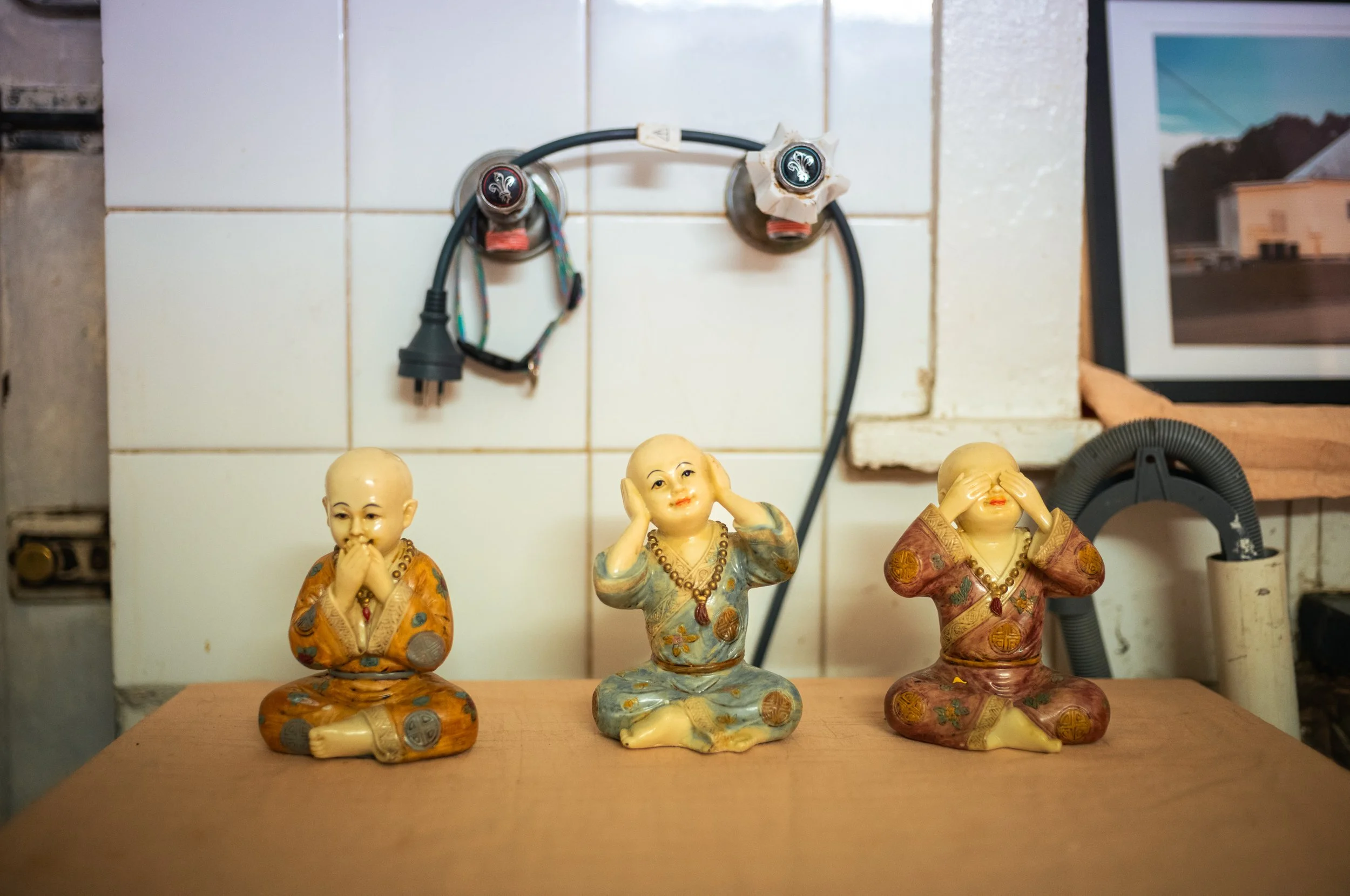 Three small Asian-style figurines sitting on a wooden surface, each covering different eyes or ears. The background has white tiled walls with electrical wires and outlets, and a framed picture partially visible on the right.