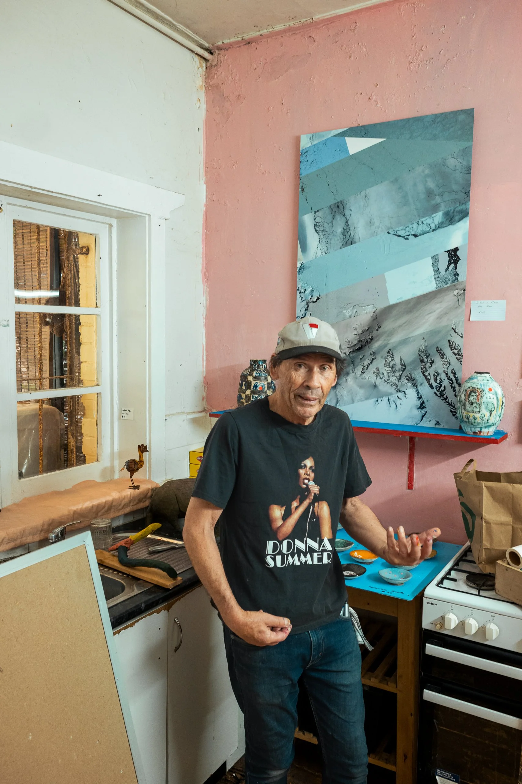 A man wearing a black Donna Summer T-shirt and a beige cap stands in a kitchen with pink walls, gesturing with his right hand. There are colorful pottery and a large abstract artwork on the wall behind him, and various kitchen items on the counter an