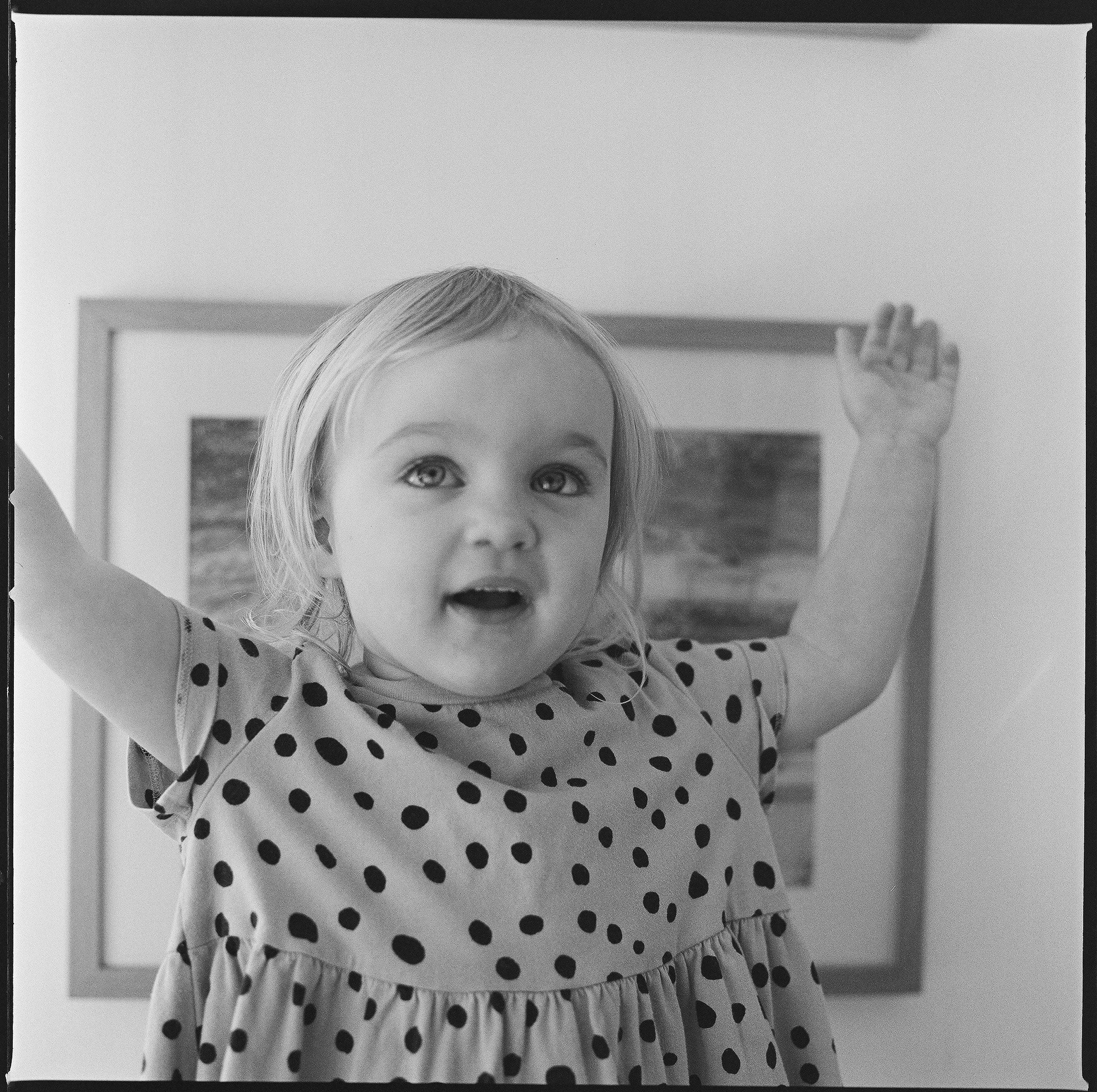 A young girl with light-colored hair, wearing a polka dot dress, standing with her arms raised and mouth open in front of a framed picture on the wall.