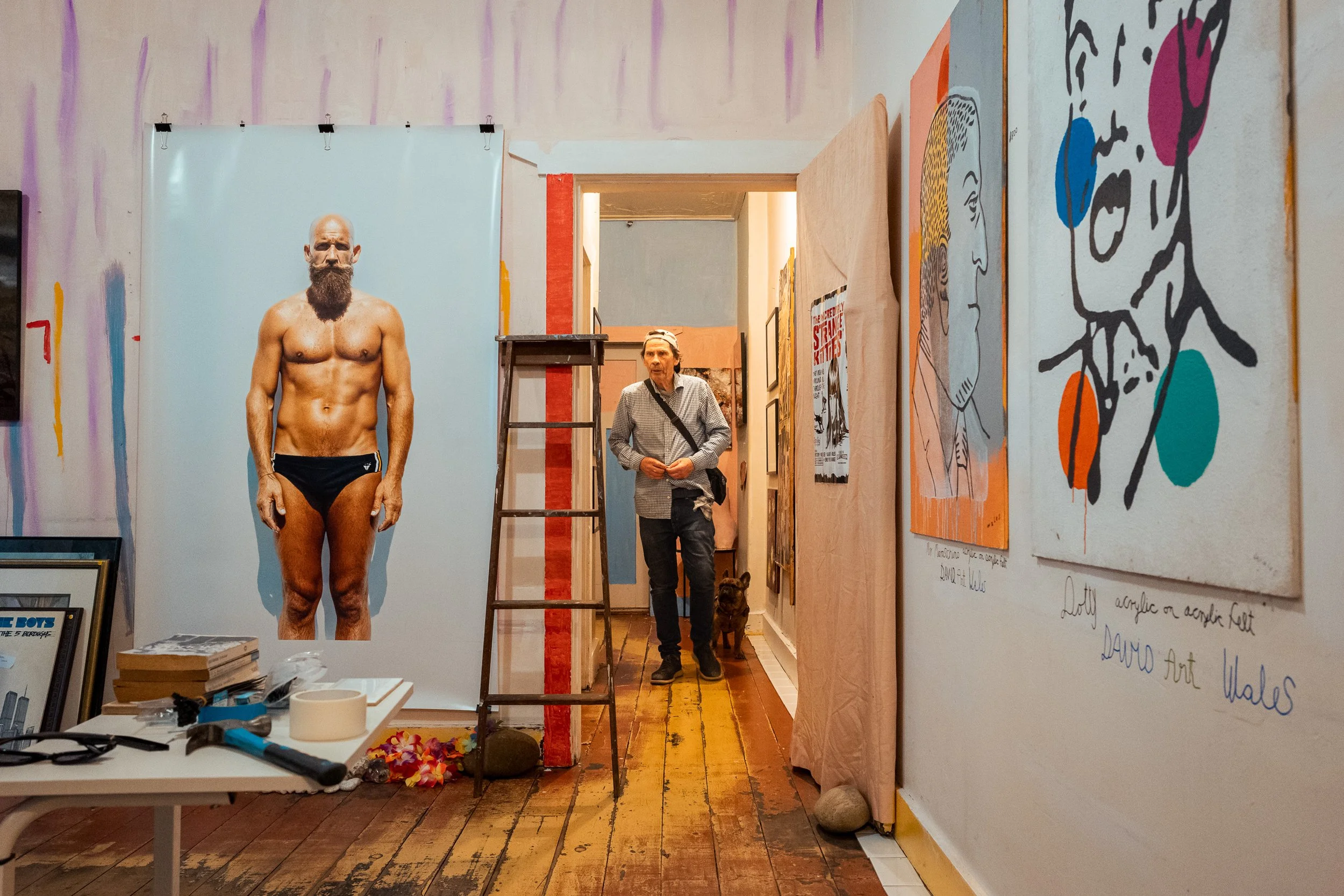 An art gallery with paintings on the walls and a shirtless, bearded man standing in front of a large photograph or painting of himself. A person with a backpack and a dog are walking down the hallway.
