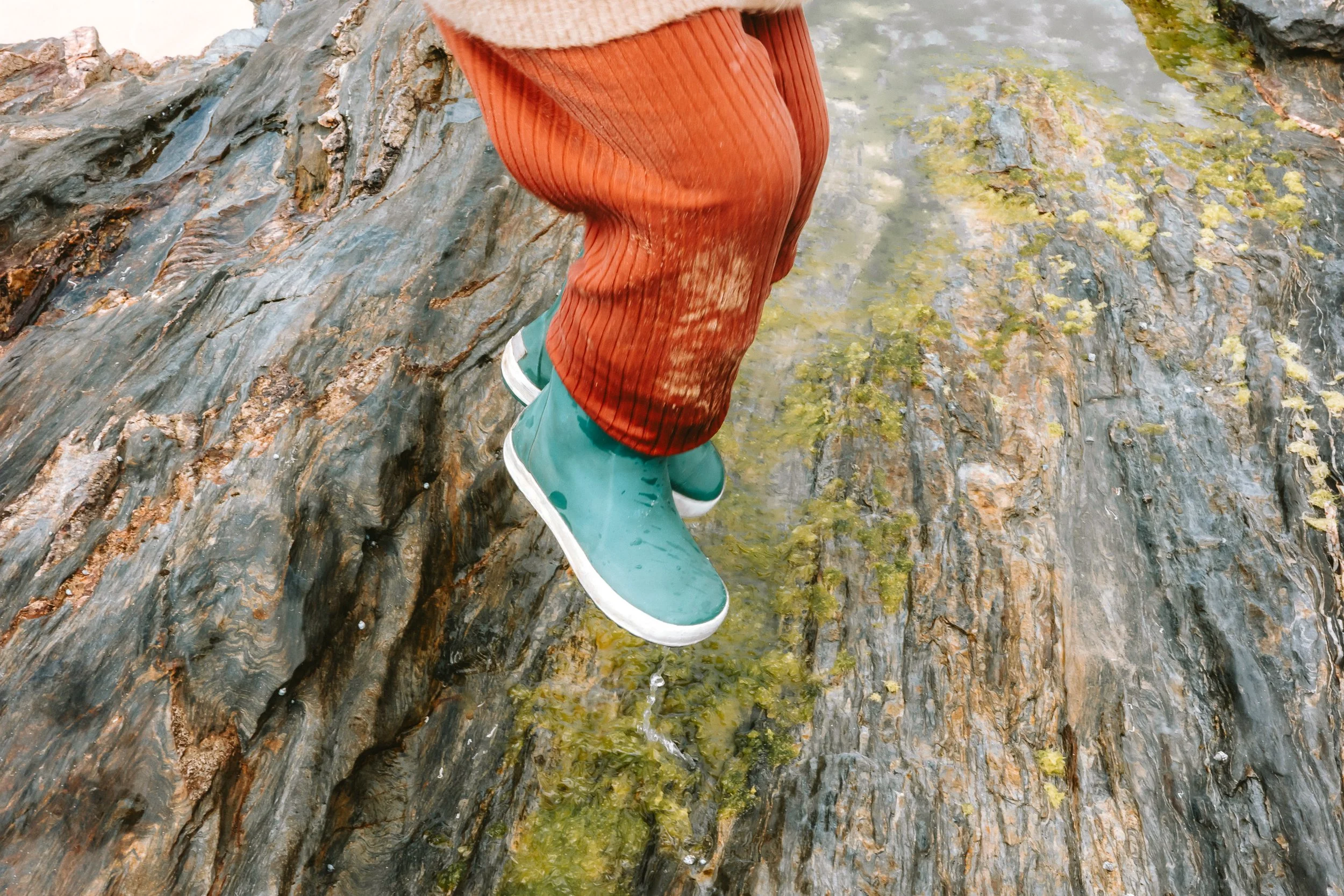 Person wearing teal rain boots and orange rain pants walking on wet rocks near water.
