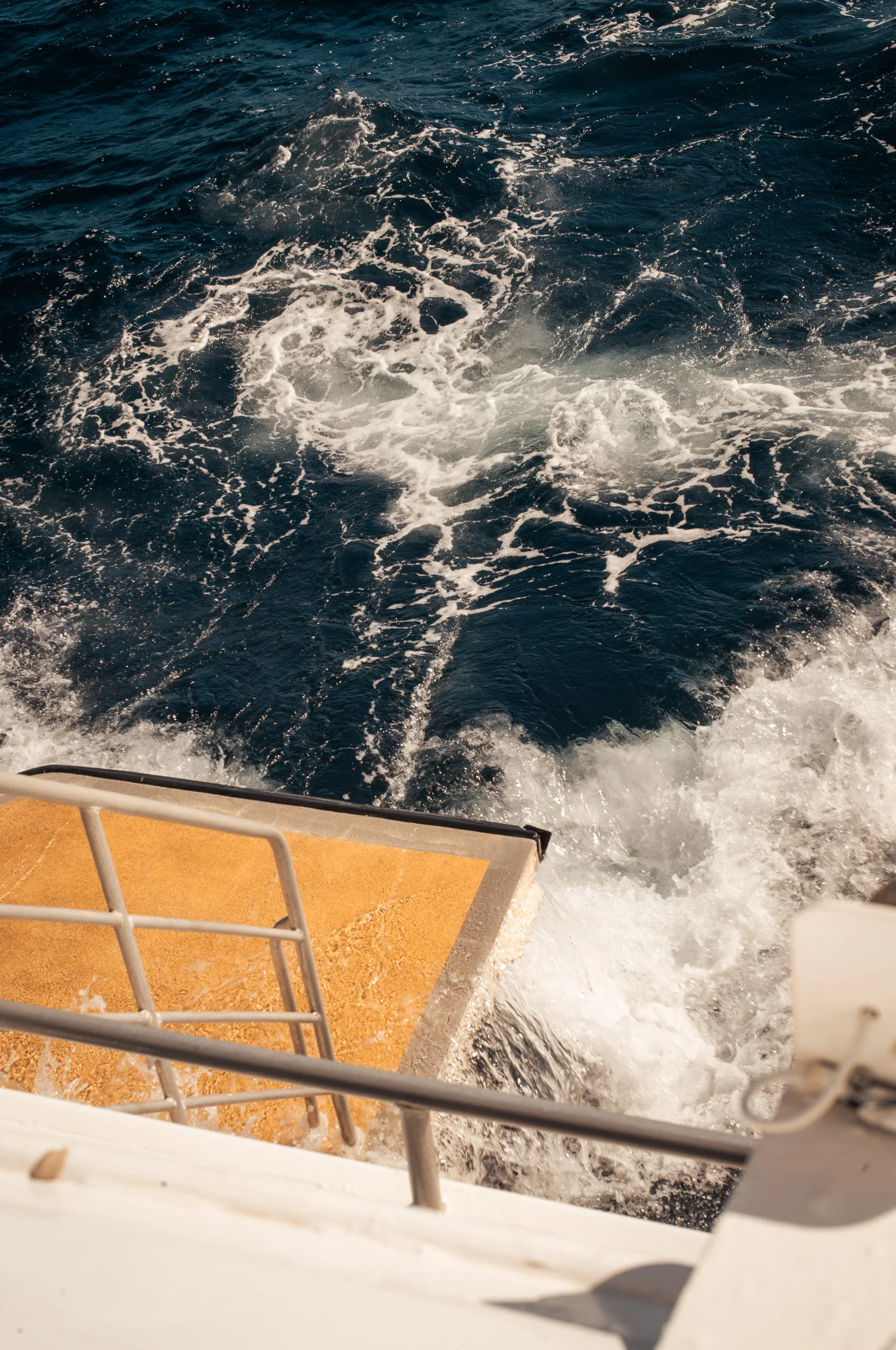 View of the ocean from a boat, showing white foamy waves and the boat's yellow deck with a metal railing in the foreground.