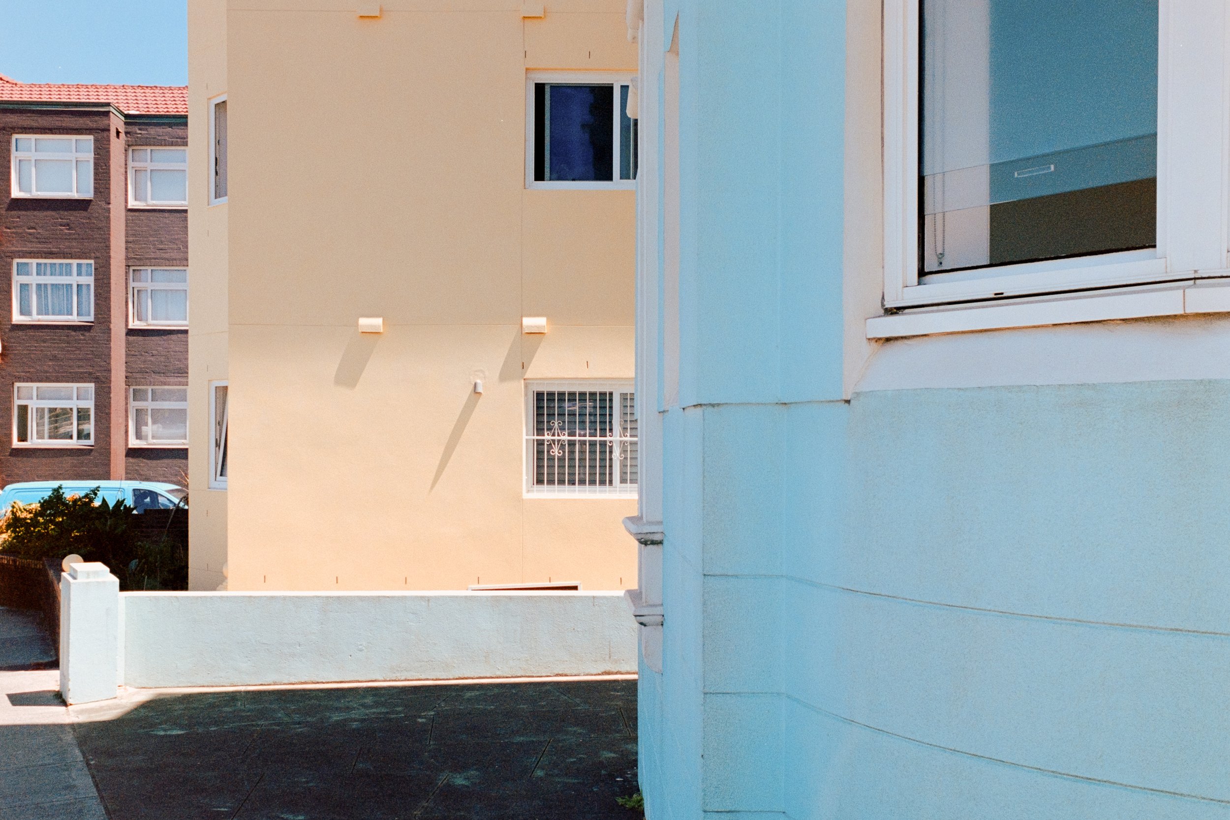 View of a light blue and beige apartment building with windows, part of a courtyard with a concrete ground, and a car parked behind a low wall.