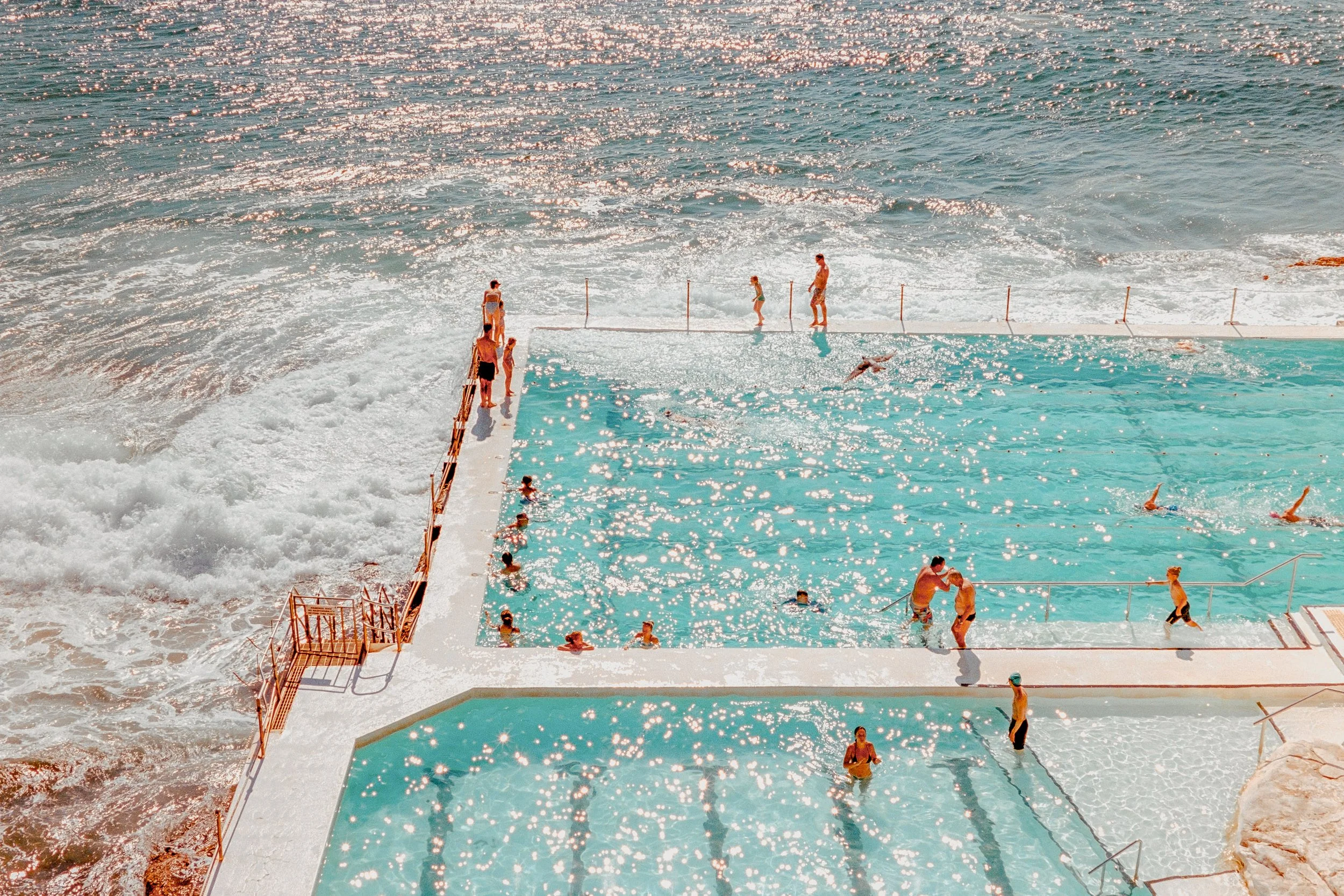 People swimming and relaxing in an oceanfront swimming pool with waves crashing nearby on a sunny day.
