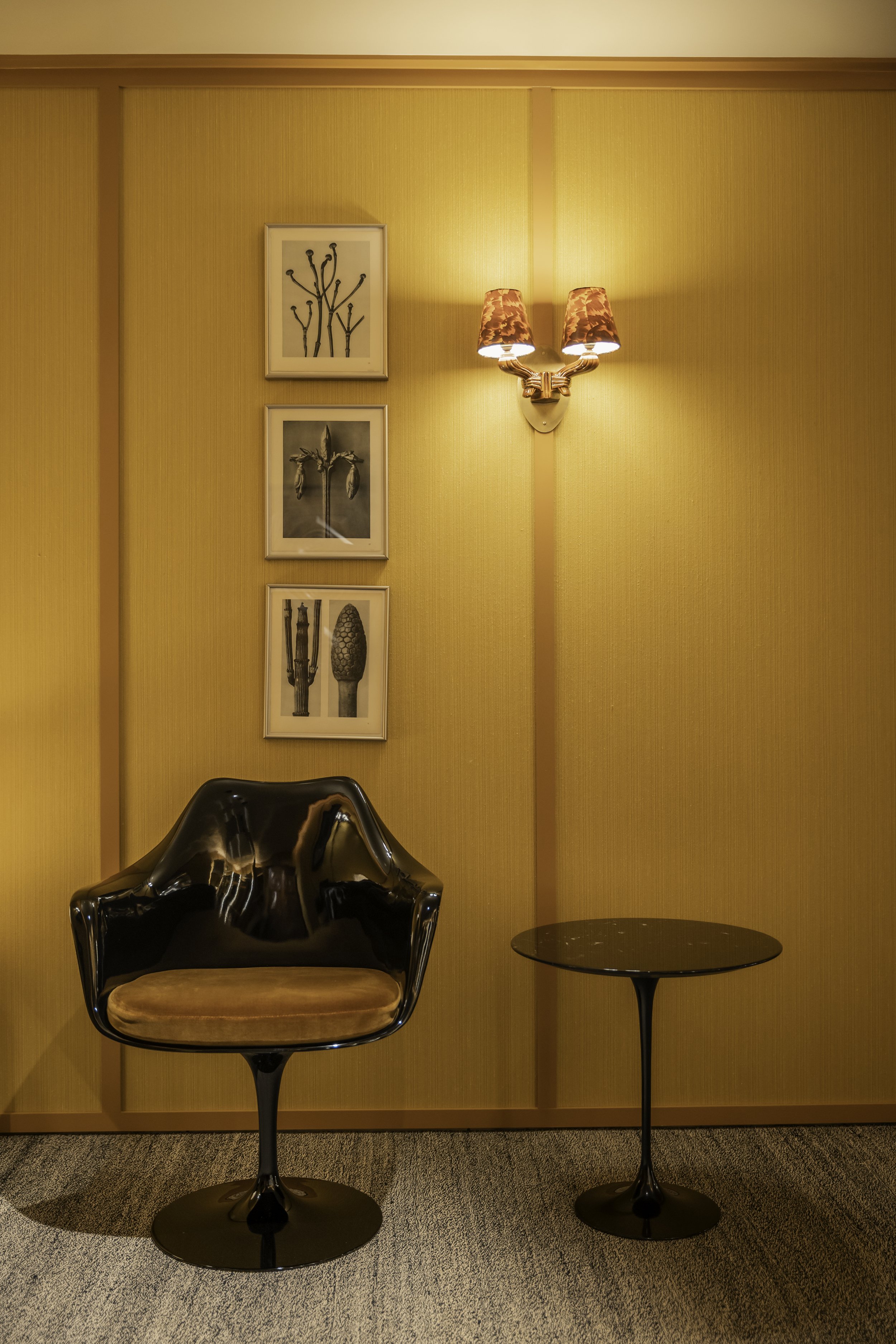A black and copper chair with a small side table on a tan carpet, yellow textured walls, framed botanical art, and a wall-mounted light fixture with two lampshades.