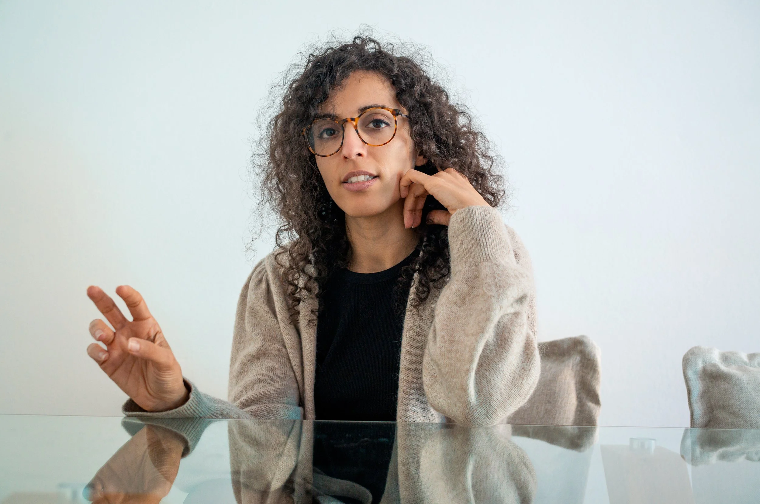 A woman with curly hair and glasses sitting at a glass table against a plain white wall, wearing a black top and beige cardigan.