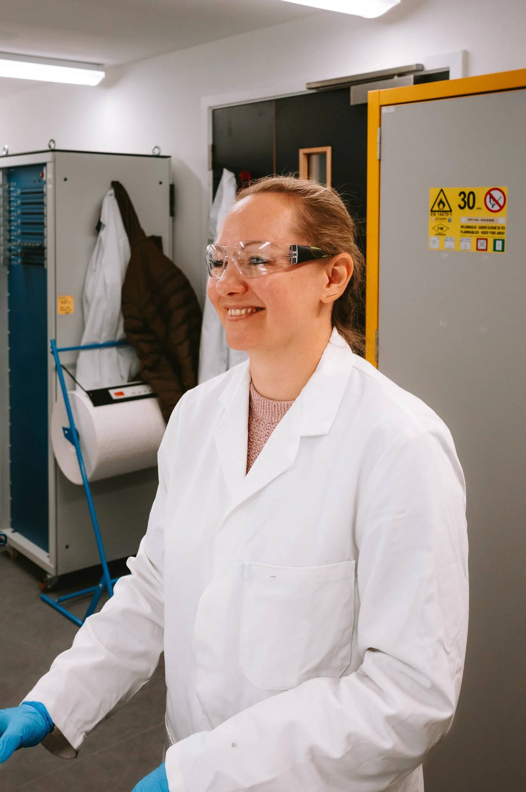 A female scientist or lab worker wearing safety goggles, a white lab coat, and blue gloves, standing in a laboratory with lockers and safety equipment in the background.