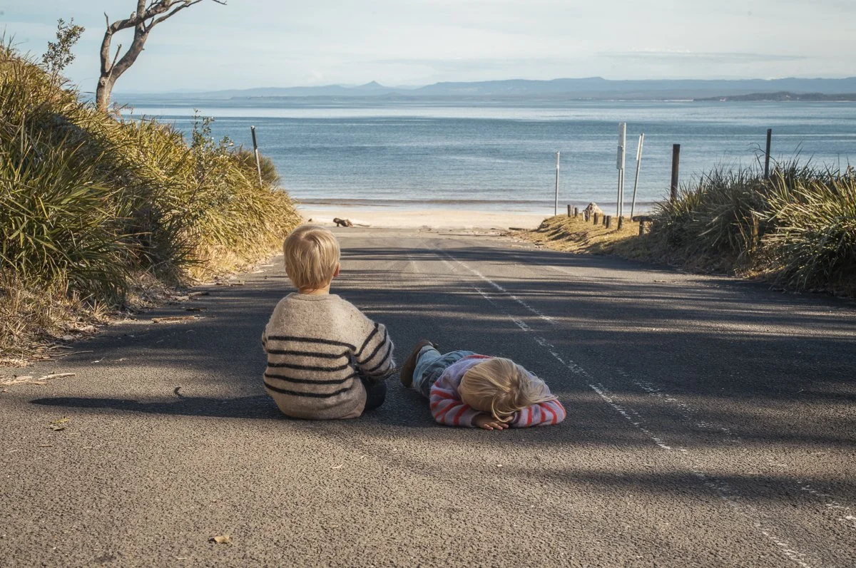 Two children, a boy and a girl, sitting on an asphalt road near a beach with water and distant mountains in the background, with the girl lying down and the boy facing away