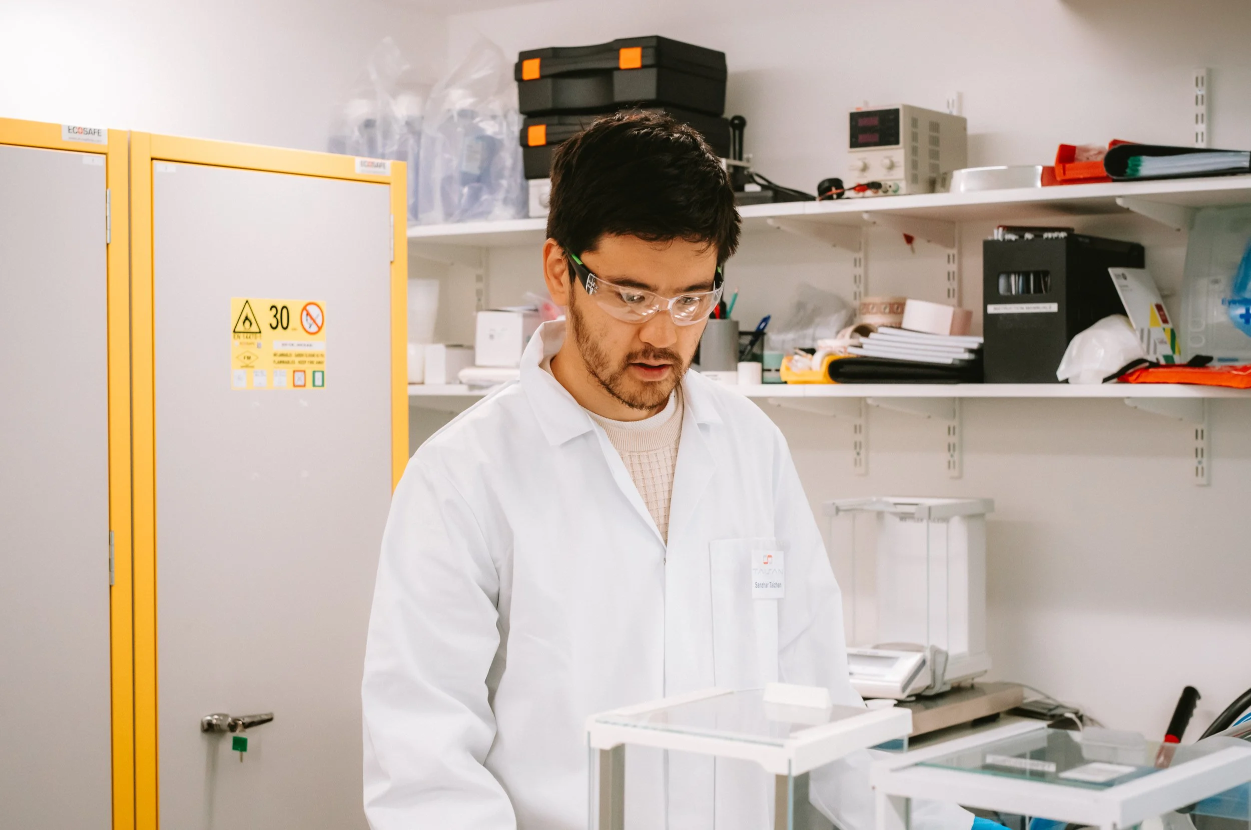 A man in a white lab coat and safety glasses working in a laboratory, surrounded by various scientific equipment and supplies on shelves.