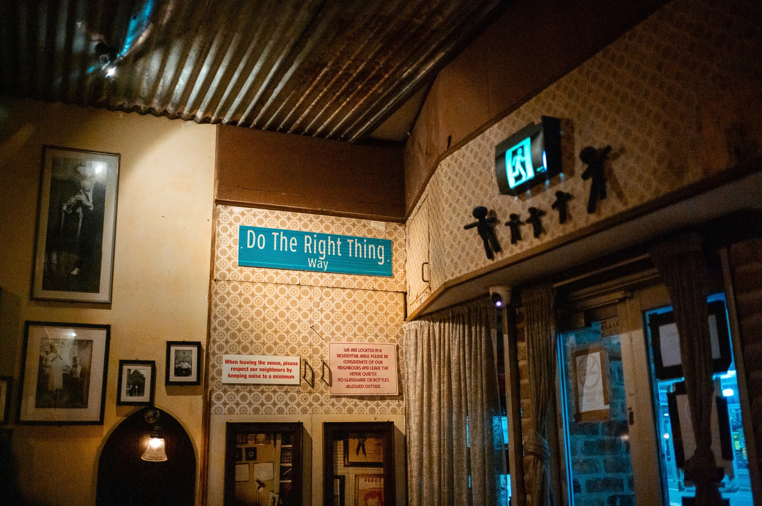 Interior of a room with vintage decor, featuring a blue sign that says 'Do The Right Thing Way,' framed photographs on the wall, a curtain covering a window or door, an emergency exit sign, and various notices and decorations.