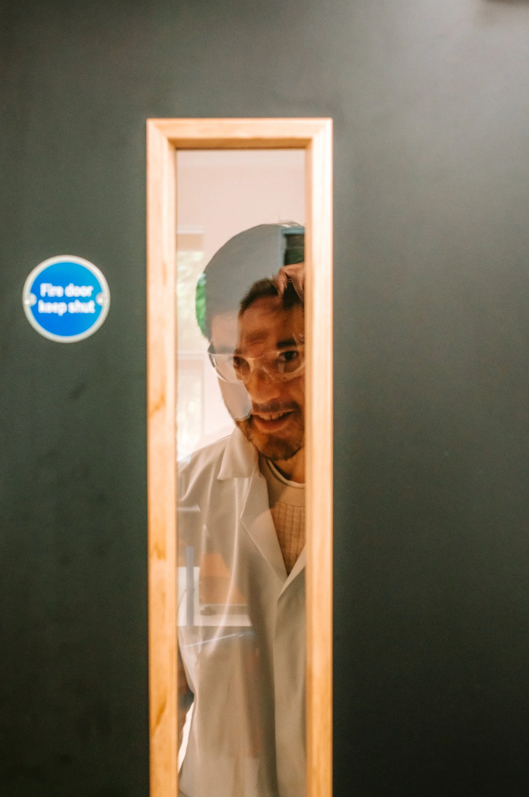 Person wearing safety goggles peeking through a small door in a black wall, with a blue sign that reads 'Fire door keep shut'.