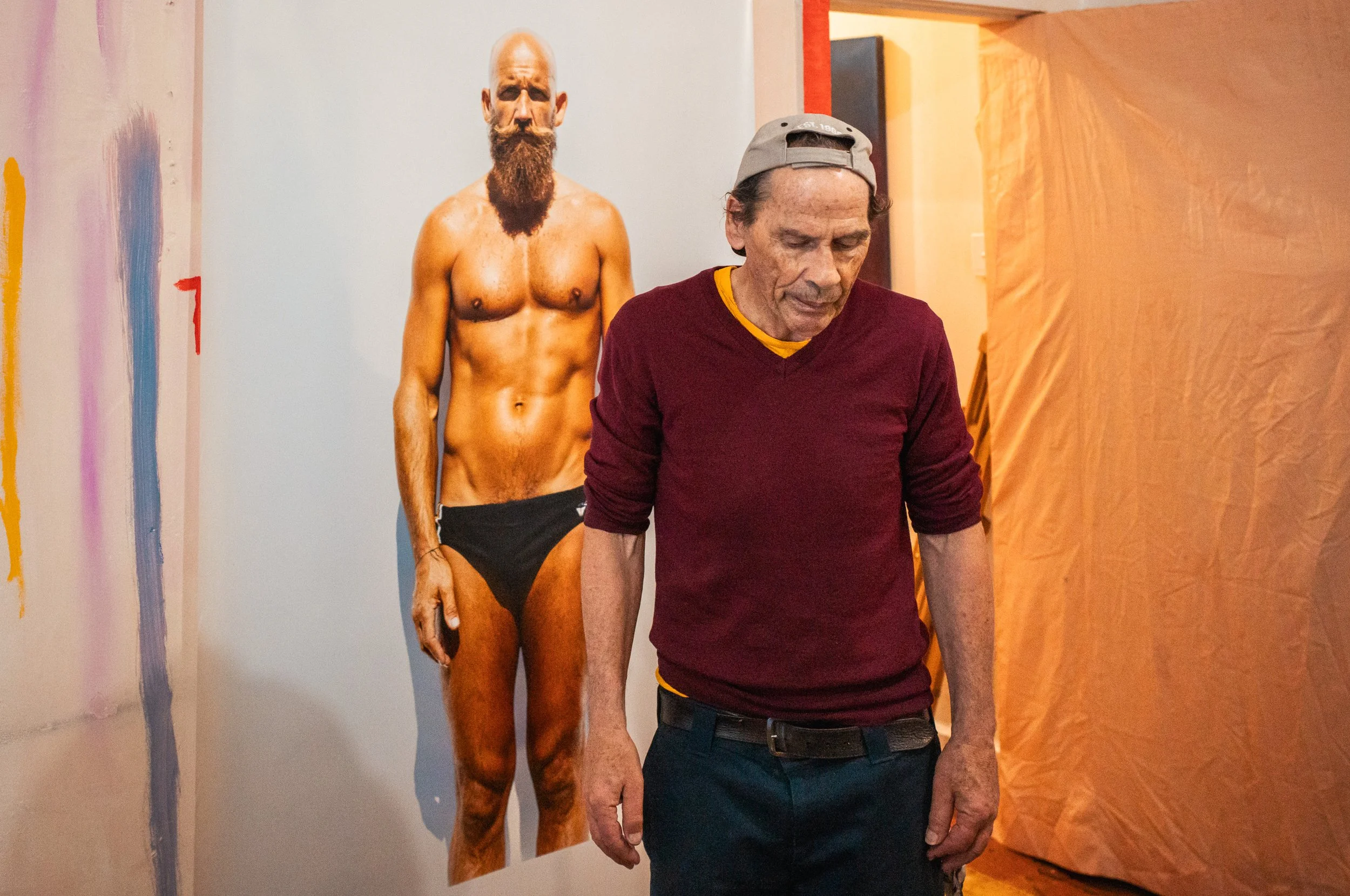 An elderly man in a maroon sweater and a backward baseball cap looks down with a solemn expression, standing in front of a painting of a shirtless man with a beard and black underwear.