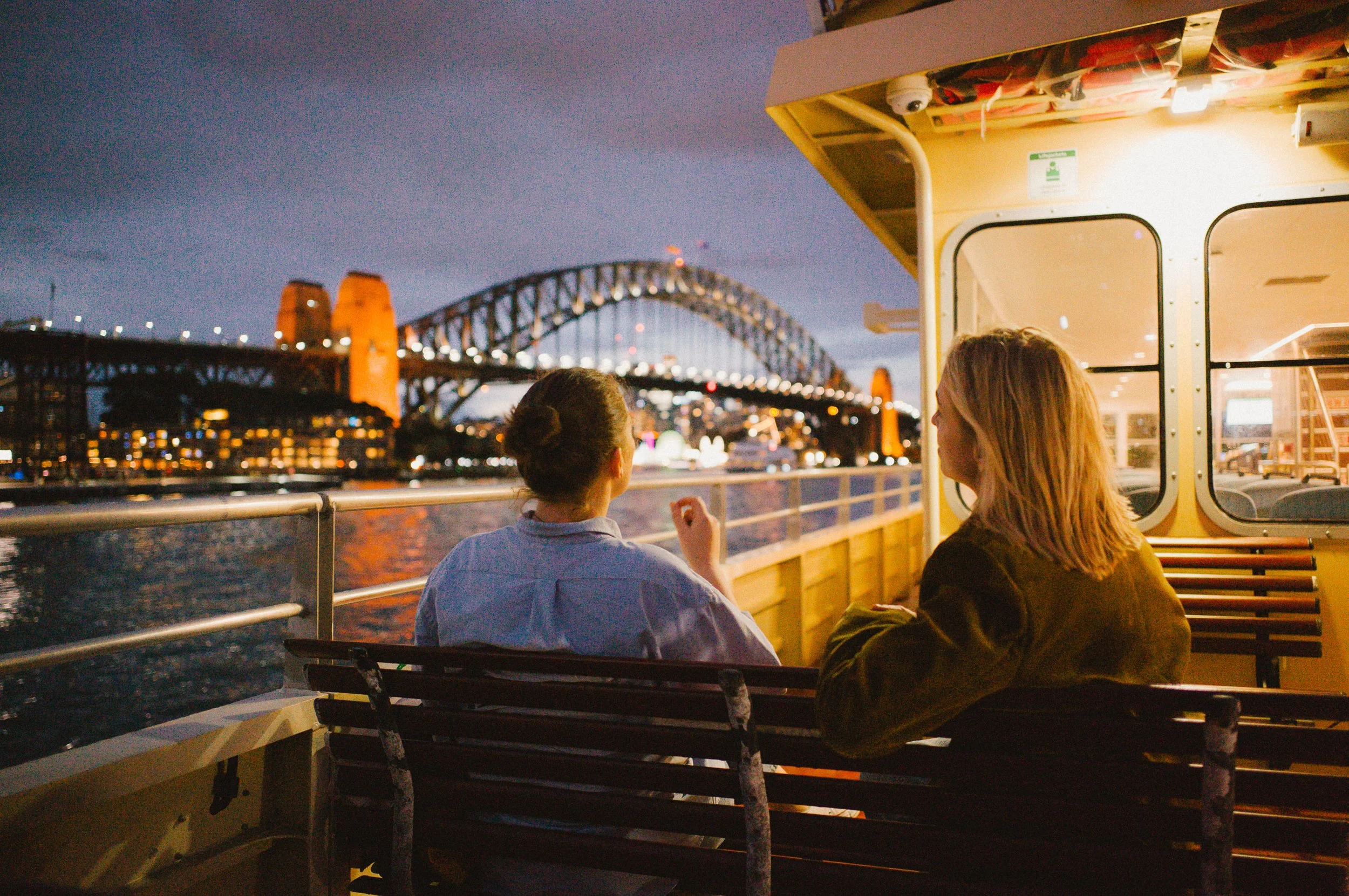 Two women seated on a boat at dusk, with the Sydney Harbour Bridge illuminated in the background.