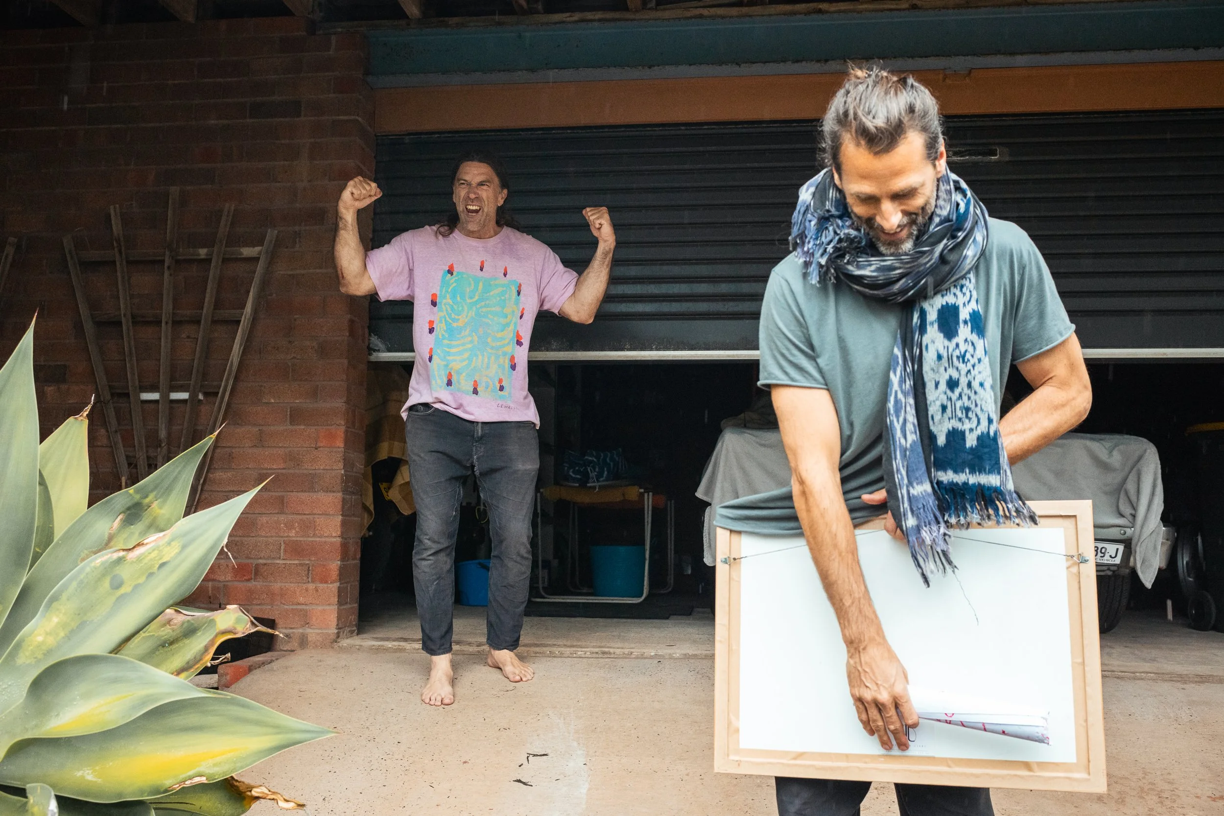 Two men in a garage celebrating, one is wearing a pink t-shirt with a birthday cake design and the other is wearing a blue scarf, holding a picture frame, and looking down.